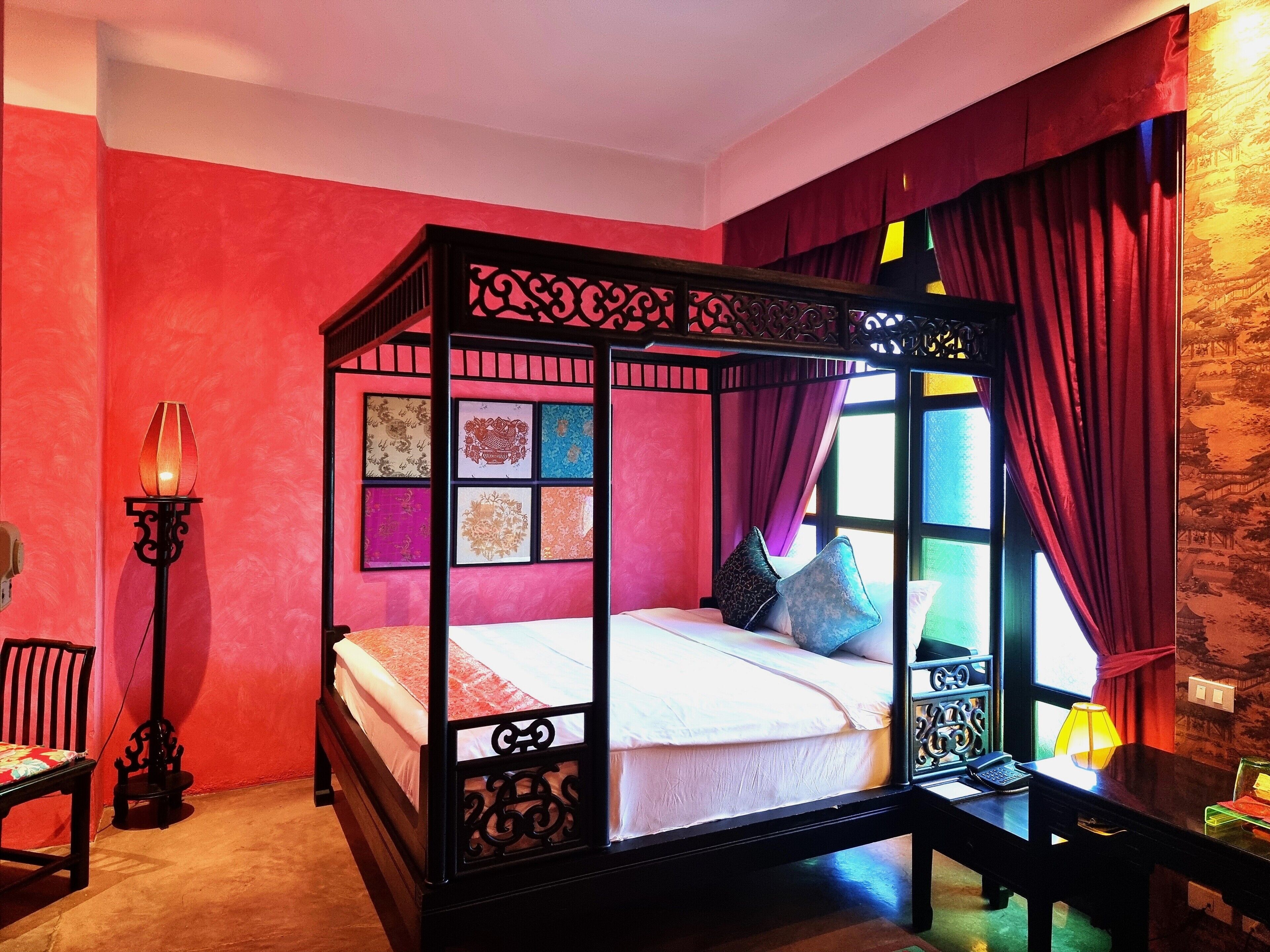 A four-poster bed with a red and black frame, a bedside table with a lamp, and a chair.