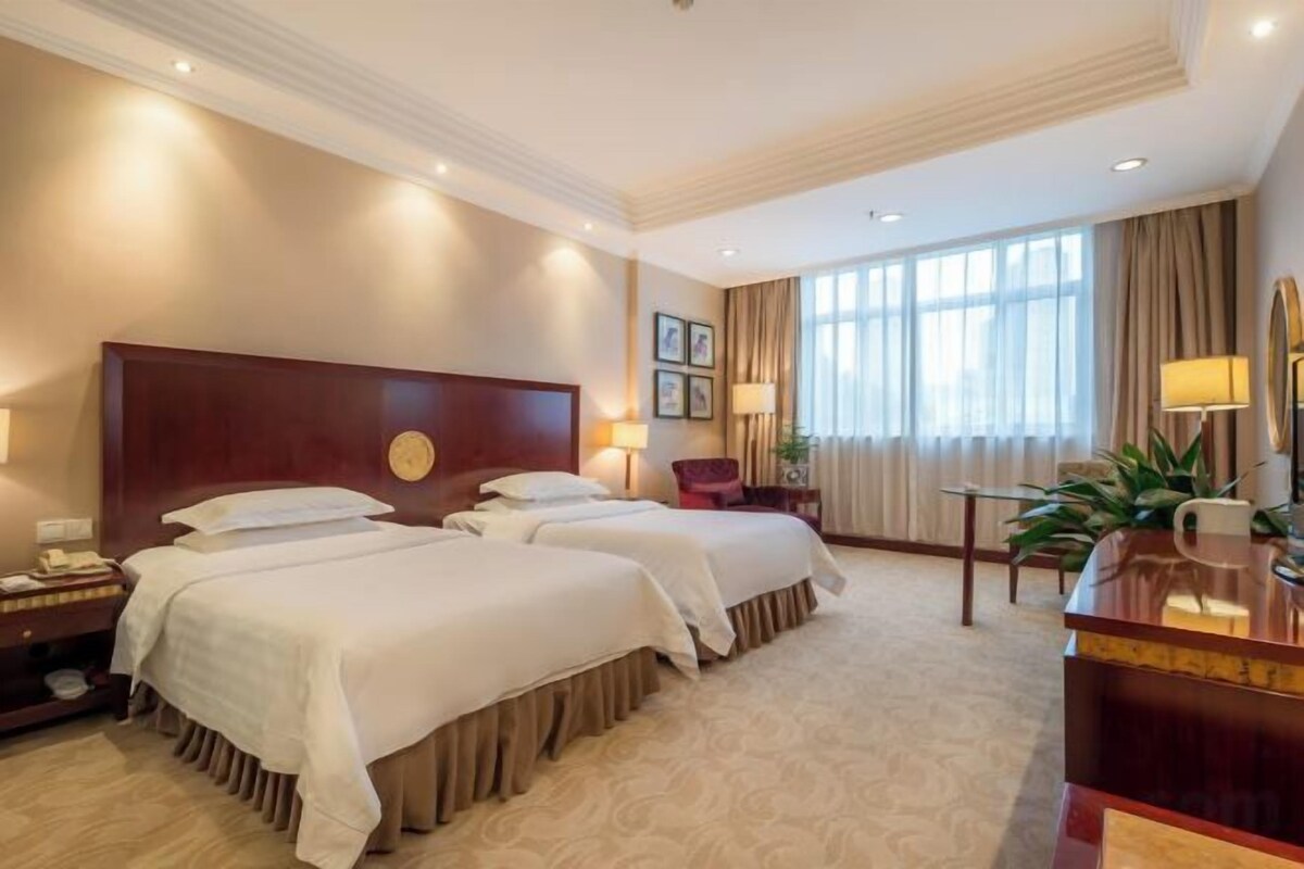 A hotel room with two beds, a desk, a chair, a nightstand, and a large window with curtains.