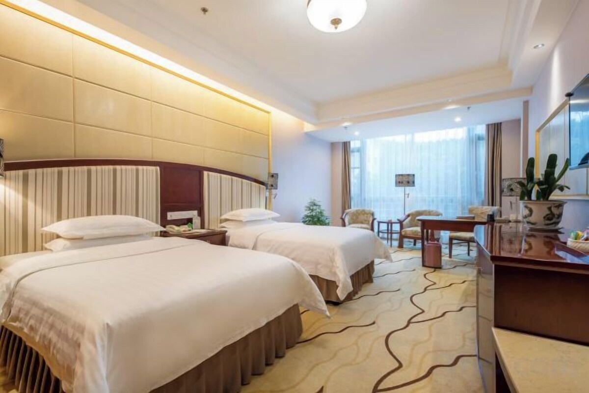 A hotel room with two beds, a desk, a chair, and a large window.