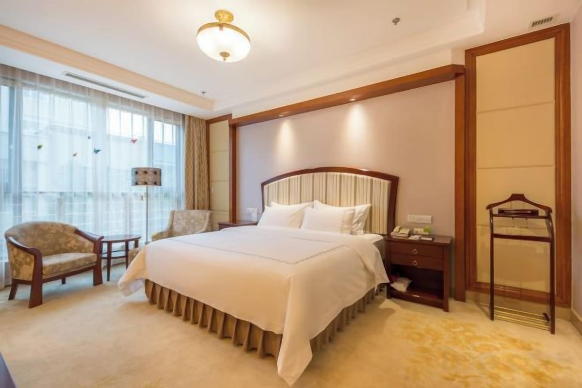 A hotel room with a large bed, a bedside table, a chair, a desk, and a wardrobe.
