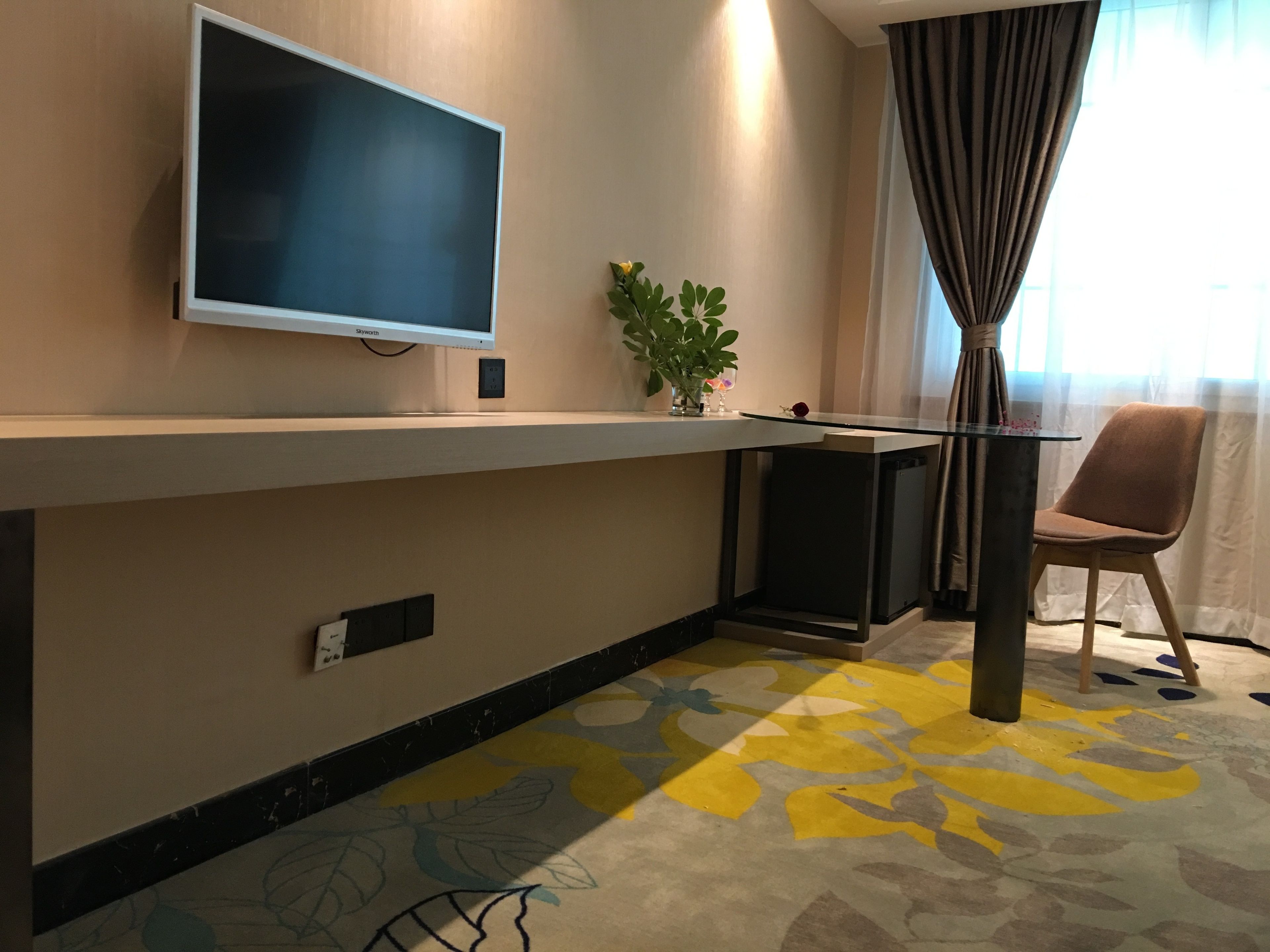 Habitación de hotel con televisión de pantalla plana montada en la pared, una mesita con planta en maceta, una silla y una alfombra colorida.