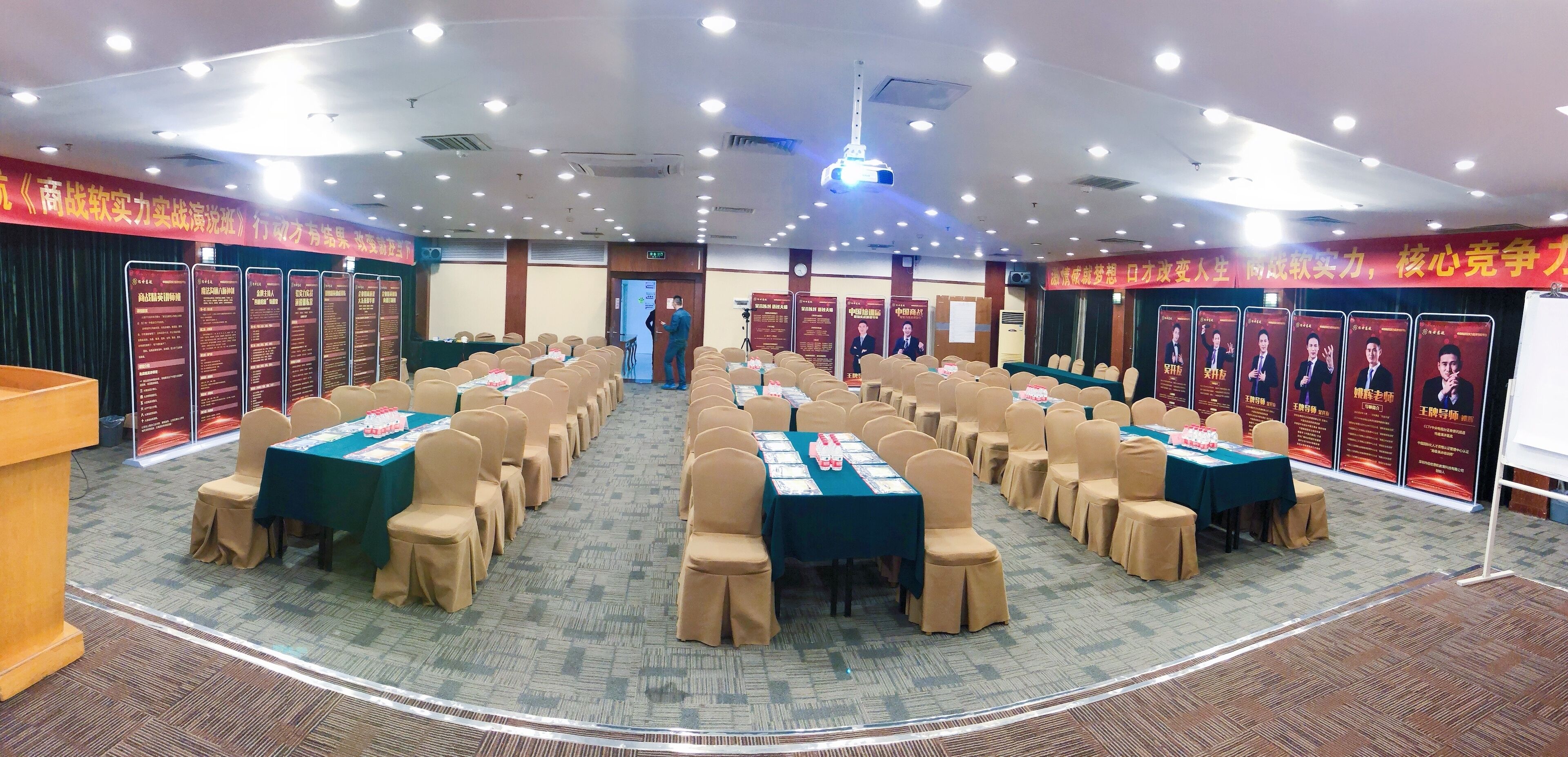 Un salón de conferencias con filas de mesas y sillas, un atril y carteles informativos en las paredes.