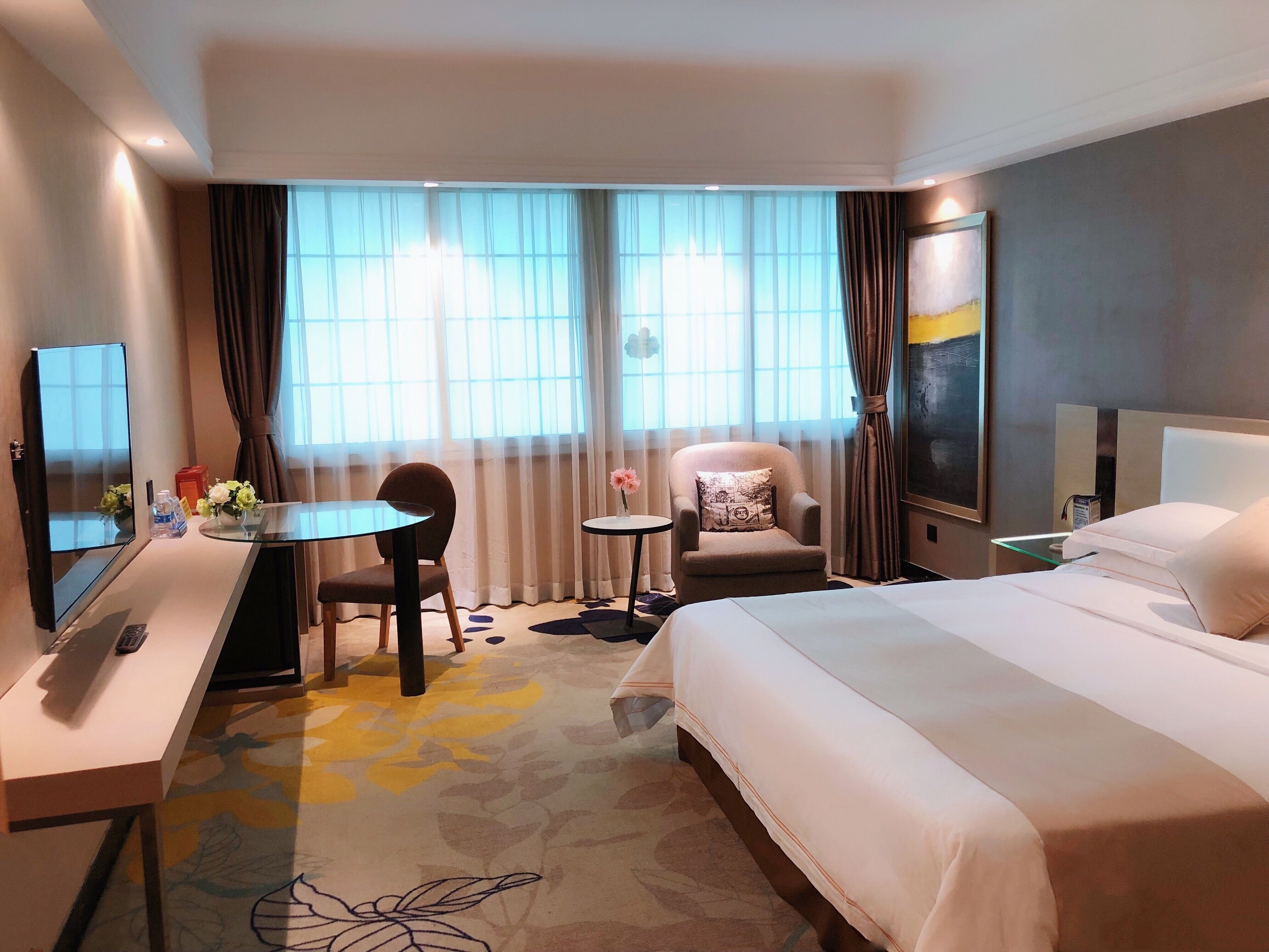 A hotel room with a bed, a desk with a computer, a chair, a small table with a vase, and a large window with curtains.