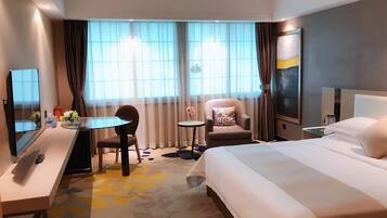 Ein Hotelzimmer mit einem Bett, einem Schreibtisch mit Computer, einem Stuhl, einem kleinen Tisch mit einer Vase und einem großen Fenster mit Vorhängen.
