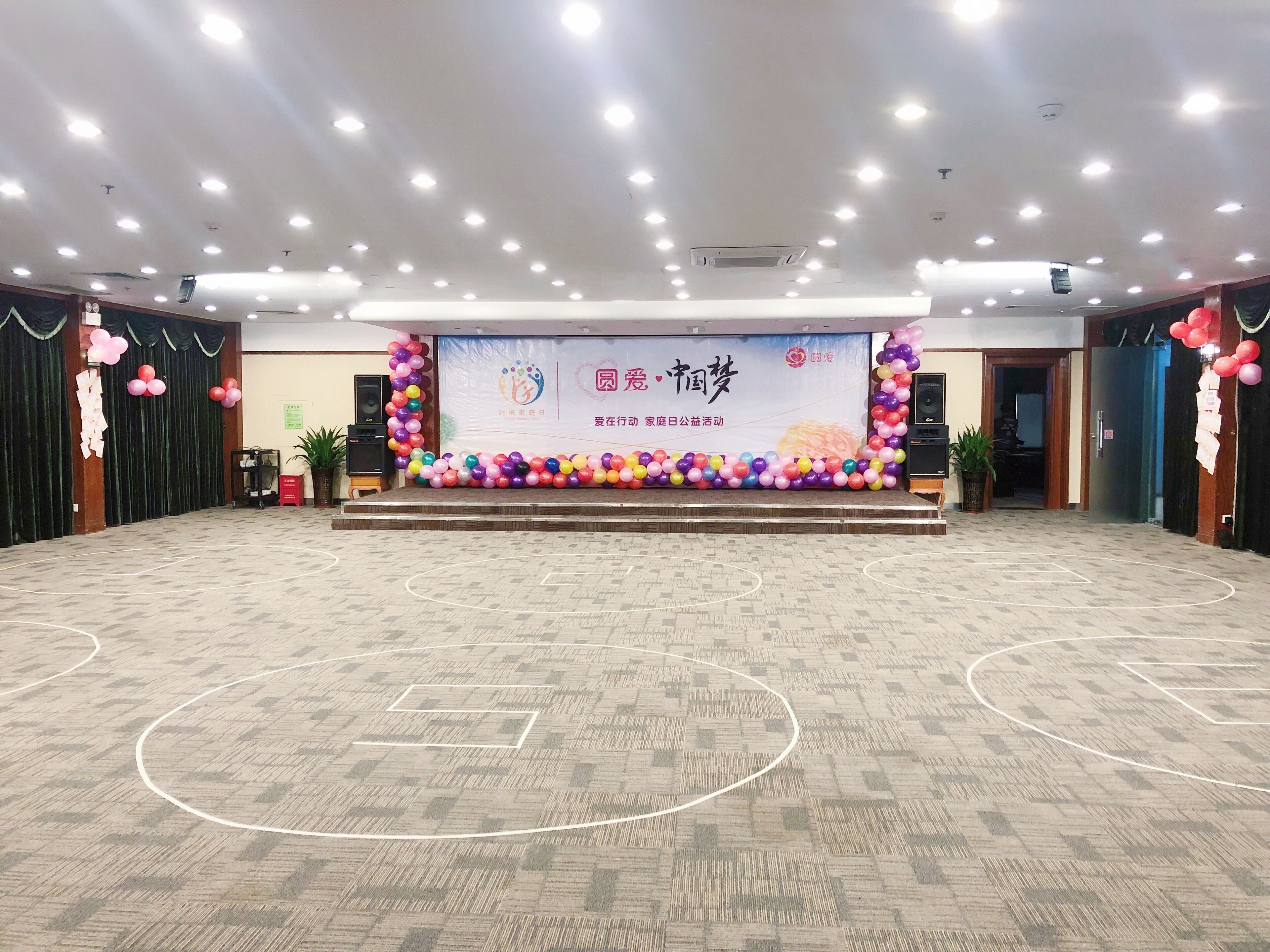 Un espacio interior amplio, con un escenario, decorado con globos de colores y un cartel.