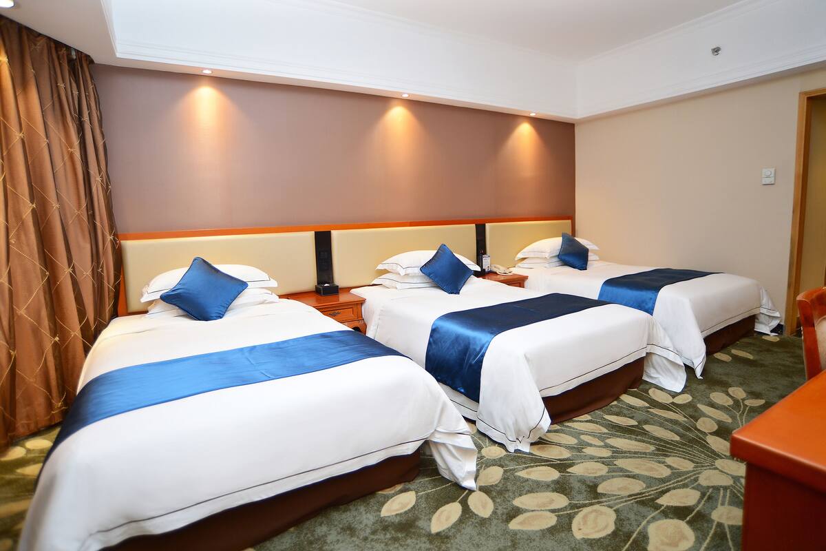 A hotel room with three beds, each with white bedding and blue accents, a patterned carpet, and a brown curtain.