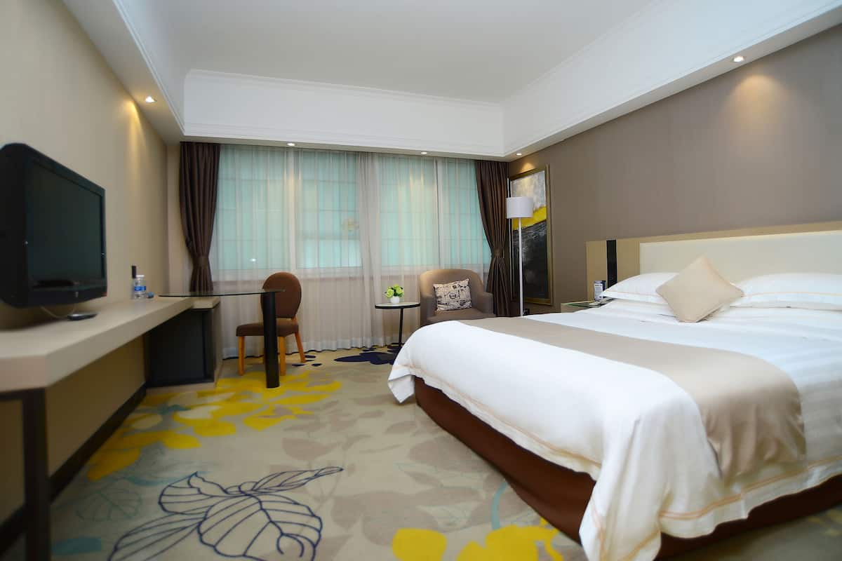 A hotel room with a large bed, a desk with a television, a chair, and a small table with a plant.