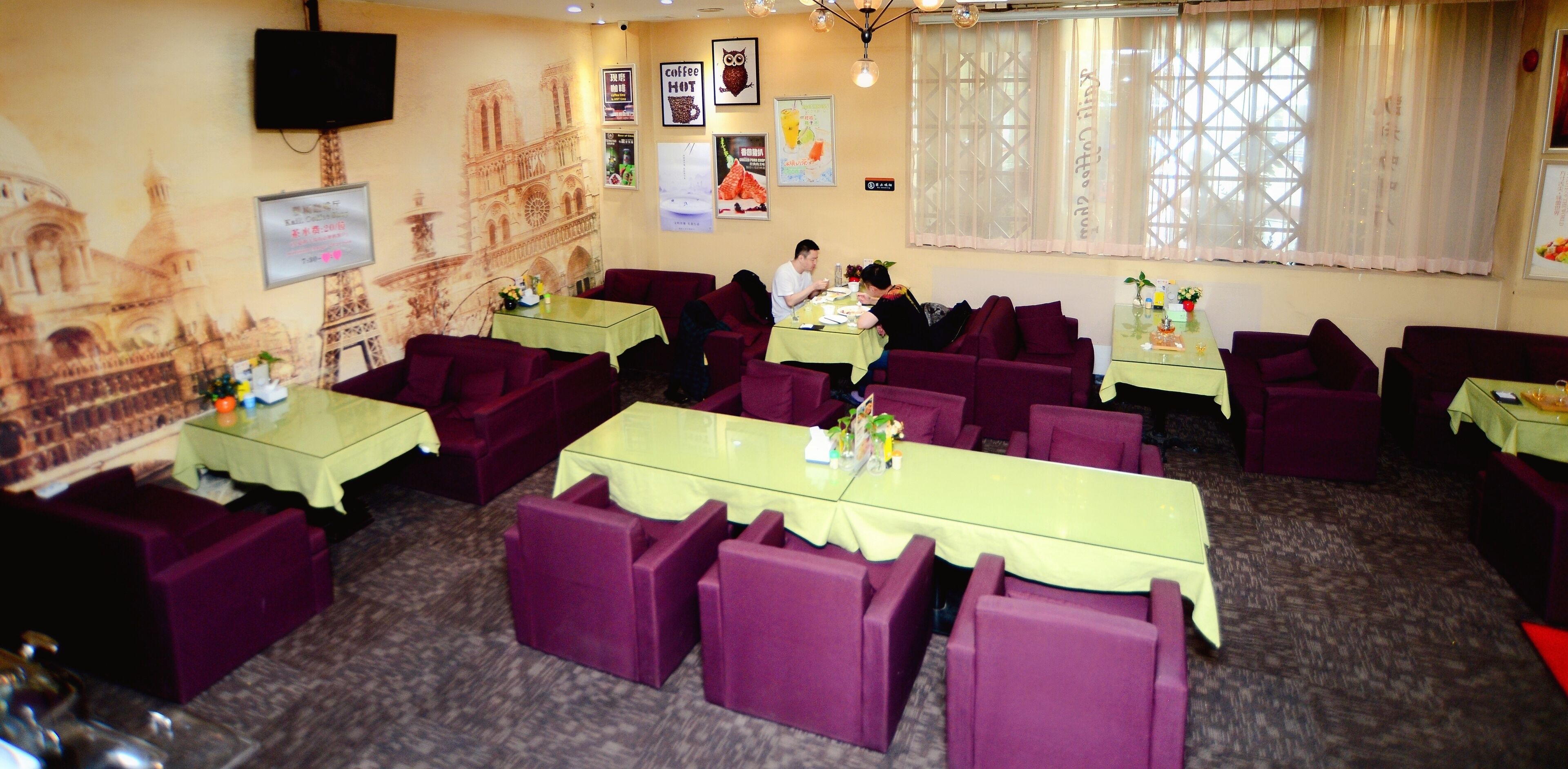 Restaurante com assentos em púrpura e mesas verdes, exibindo um mural de um edifício famoso e diversos cartazes nas paredes.