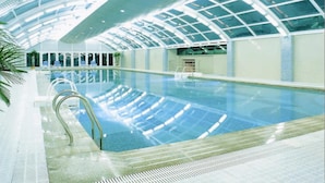 Una piscina interior con techo de vidrio, agua cristalina y suelo de baldosas.