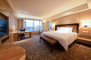 Habitación de hotel con una cama grande, un escritorio con computadora, televisor y vista a la ciudad.
