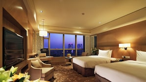 Habitación de hotel con televisor grande, dos camas, un escritorio, una silla, una mesita con flores y vista a la ciudad al anochecer.