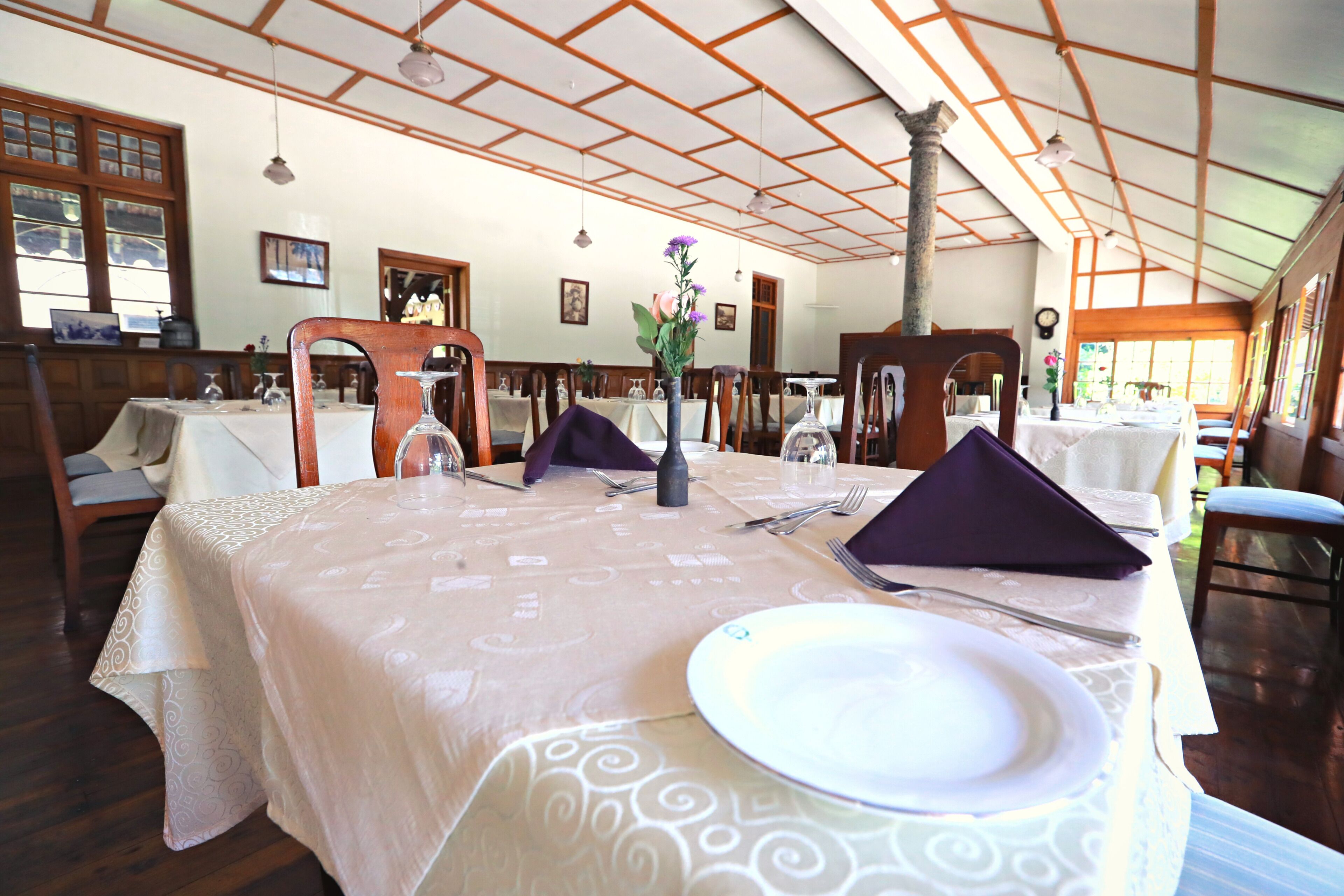 A dining area with tables set for two, wooden chairs, and a high ceiling with wooden beams.