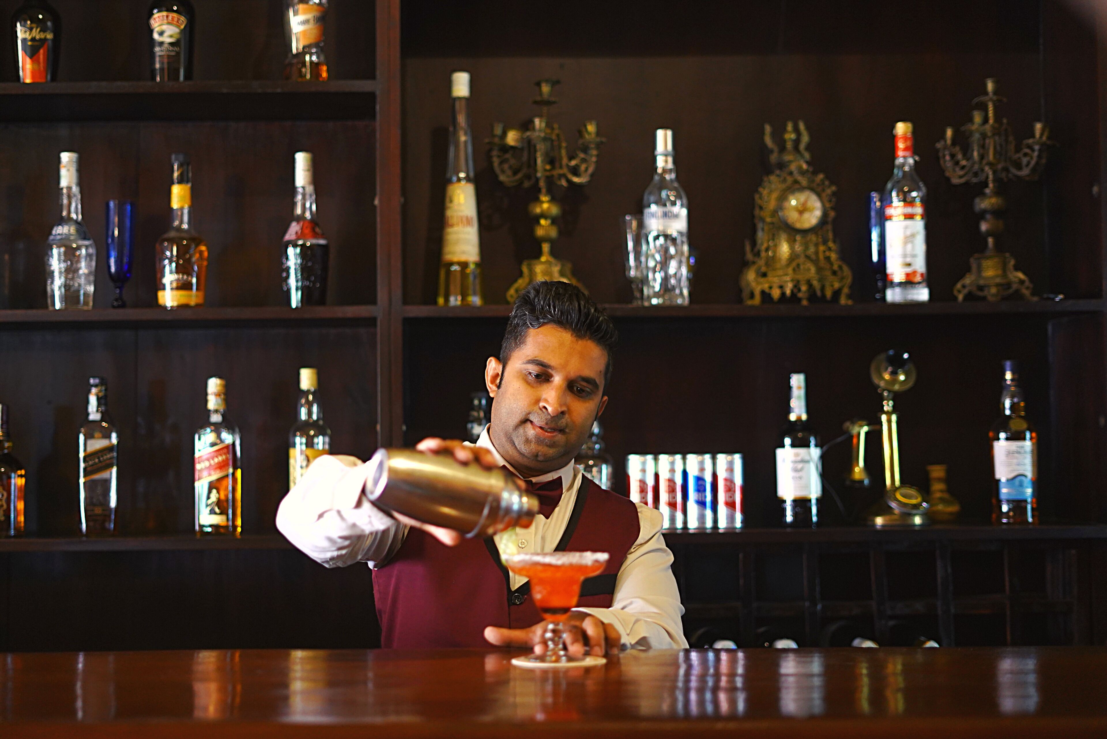 A bartender pouring a drink at a bar with shelves of liquor bottles and ornate chandeliers in the background.