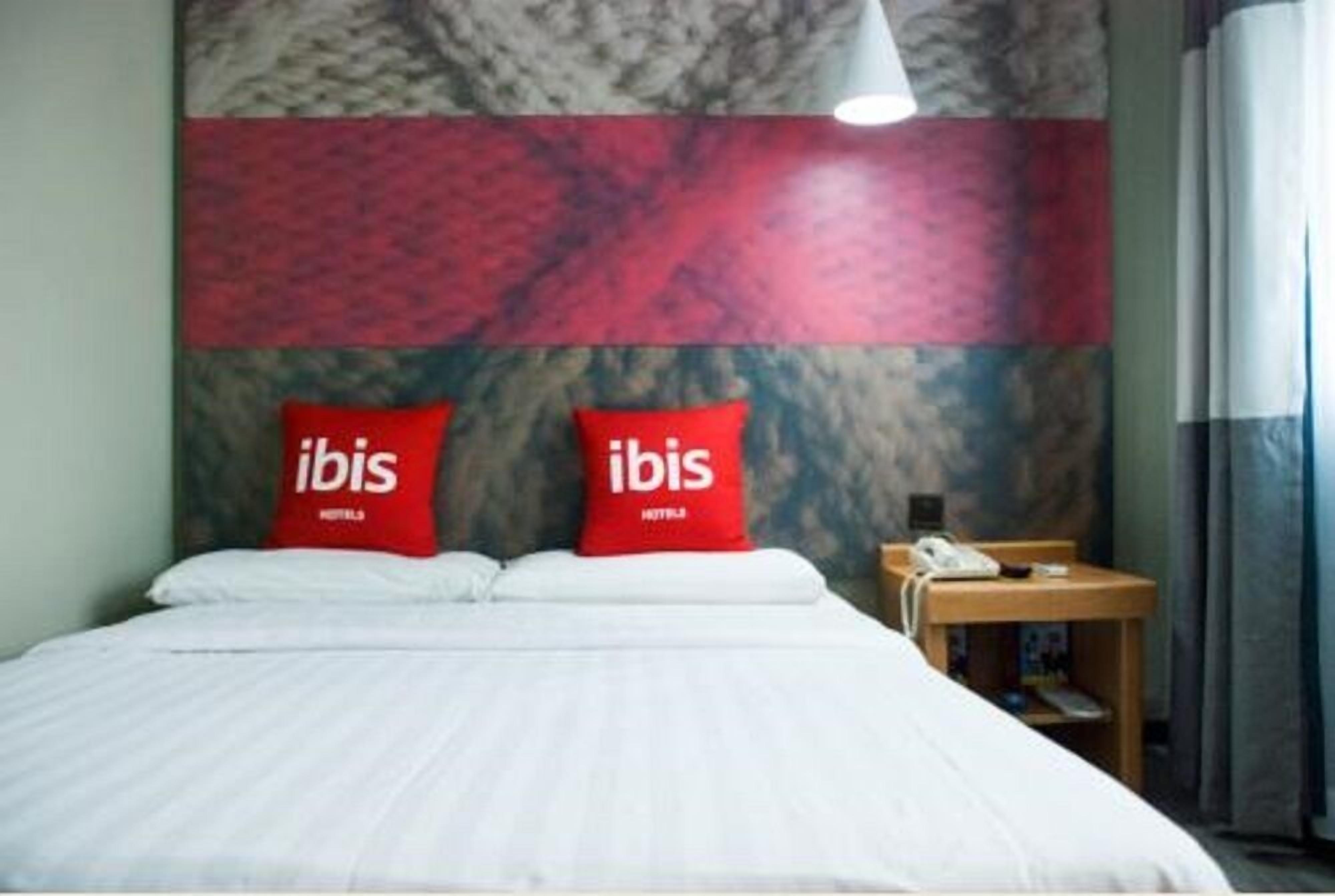 A hotel room with a bed, two red pillows with 'ibis' branding, a marble-patterned wall, a bedside table with a phone, and a lamp.