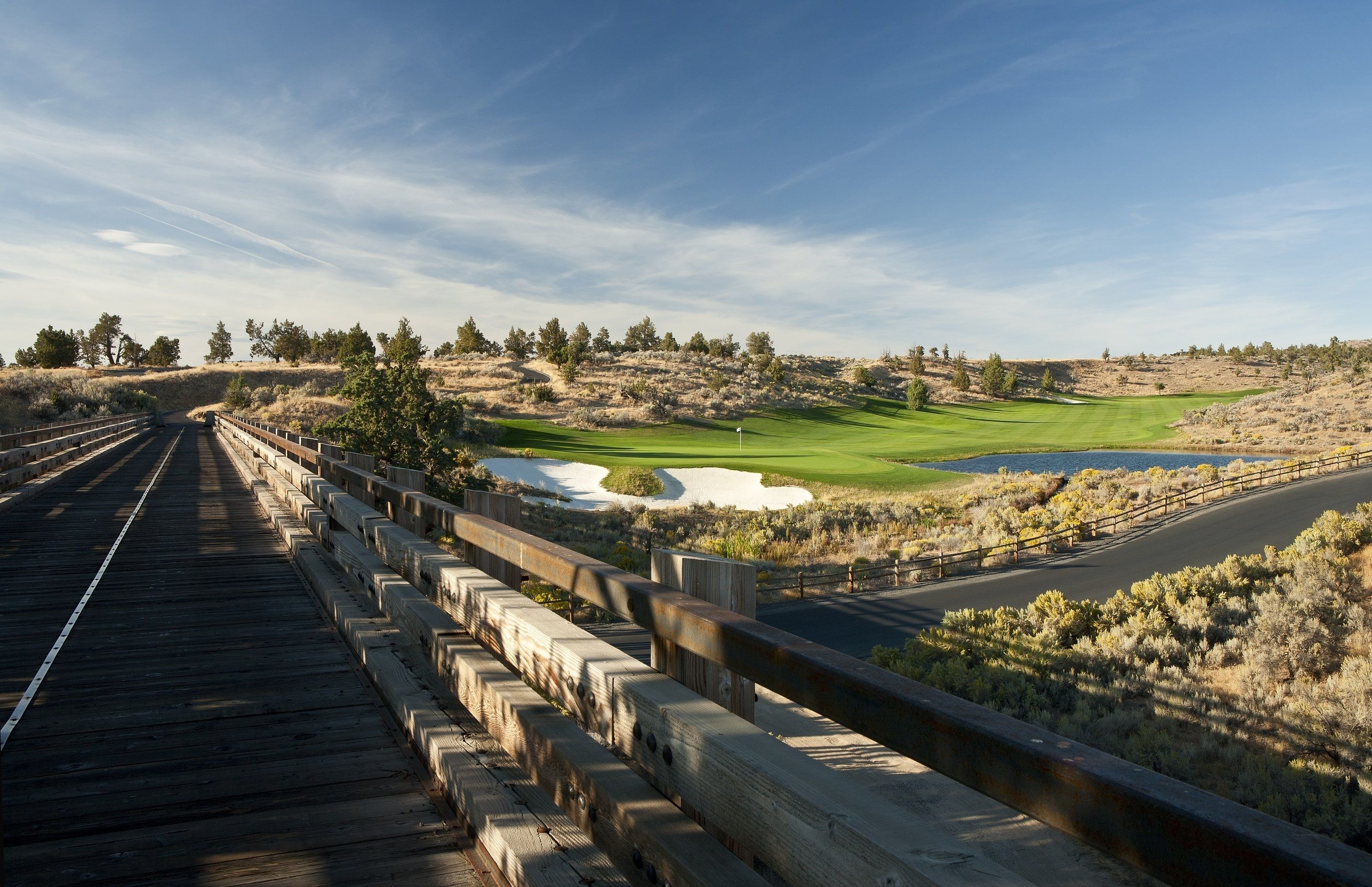 Un puente de madera con barandilla que conduce a un campo de golf con un estanque y búnkeres de arena.