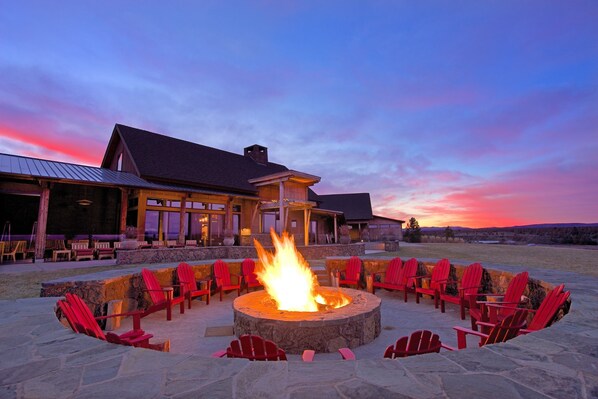 A stone fire pit with red chairs arranged around it, set against a backdrop of a building with large windows and a sloped roof.