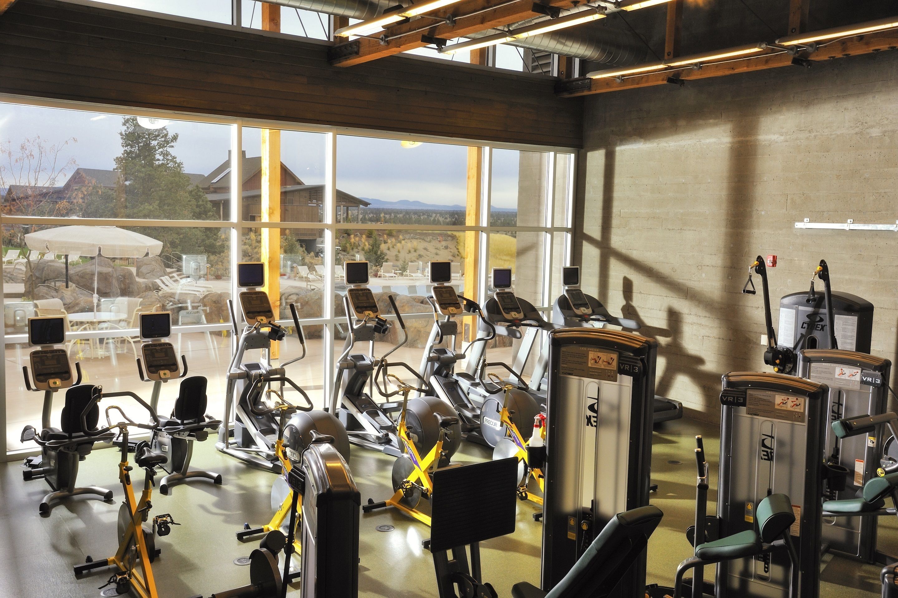 Ein Fitnessstudio mit verschiedenen Trainingsgeräten, großen Fenstern und Blick auf Sitzmöglichkeiten im Freien sowie auf Berge.