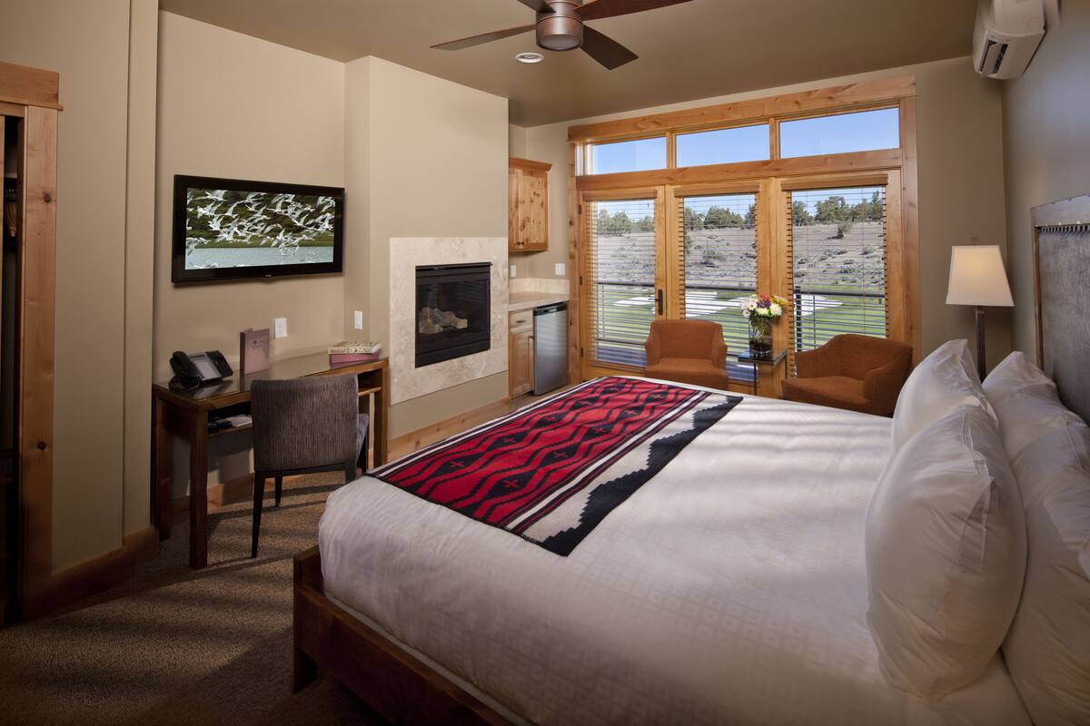A bedroom with a large bed, a desk, a chair, a fireplace, and a view of the outdoors.