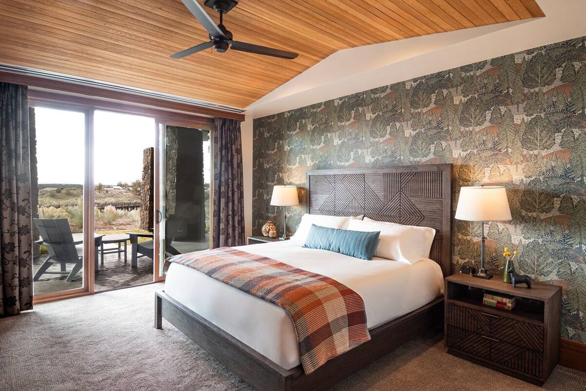 A bedroom with a large bed, two bedside lamps, a wooden headboard, and a view of the outdoors through a sliding glass door.