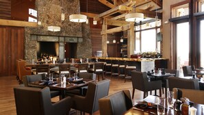 A spacious restaurant with high ceilings, exposed wooden beams, stone walls, and a central fireplace. Tables and chairs are arranged for dining, and a bar area is visible in the background.