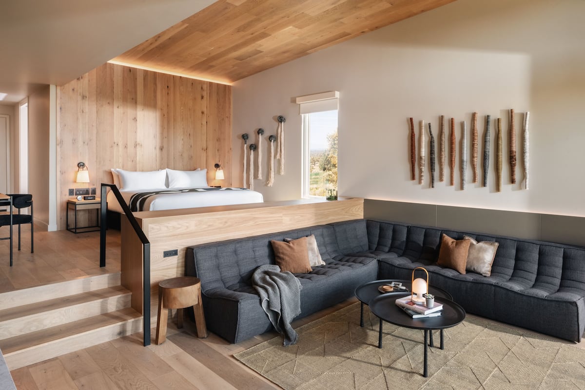 A modern hotel room with a loft bed, a dark grey sofa, a small coffee table, and a large window.