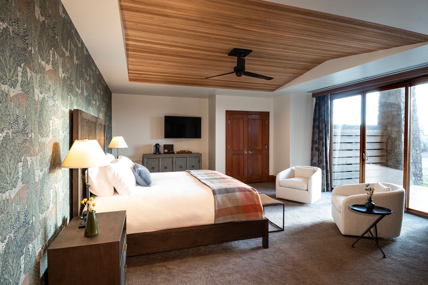 A modern hotel room with a large bed, a desk, a chair, a television, and a balcony with a view.