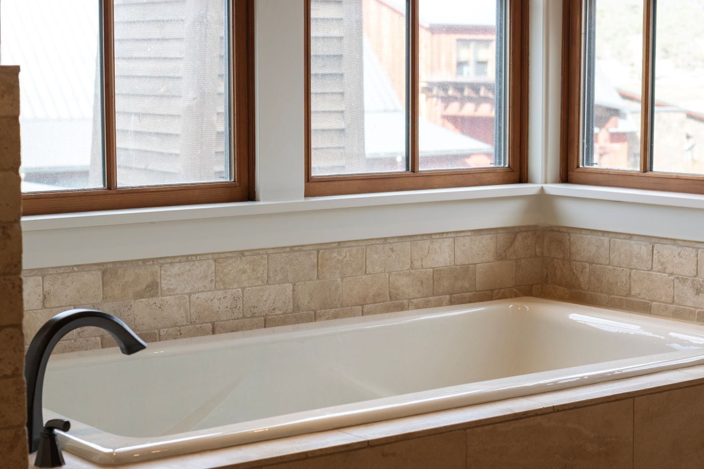 A bathroom with a white bathtub, beige tiles, and a window with a wooden frame.