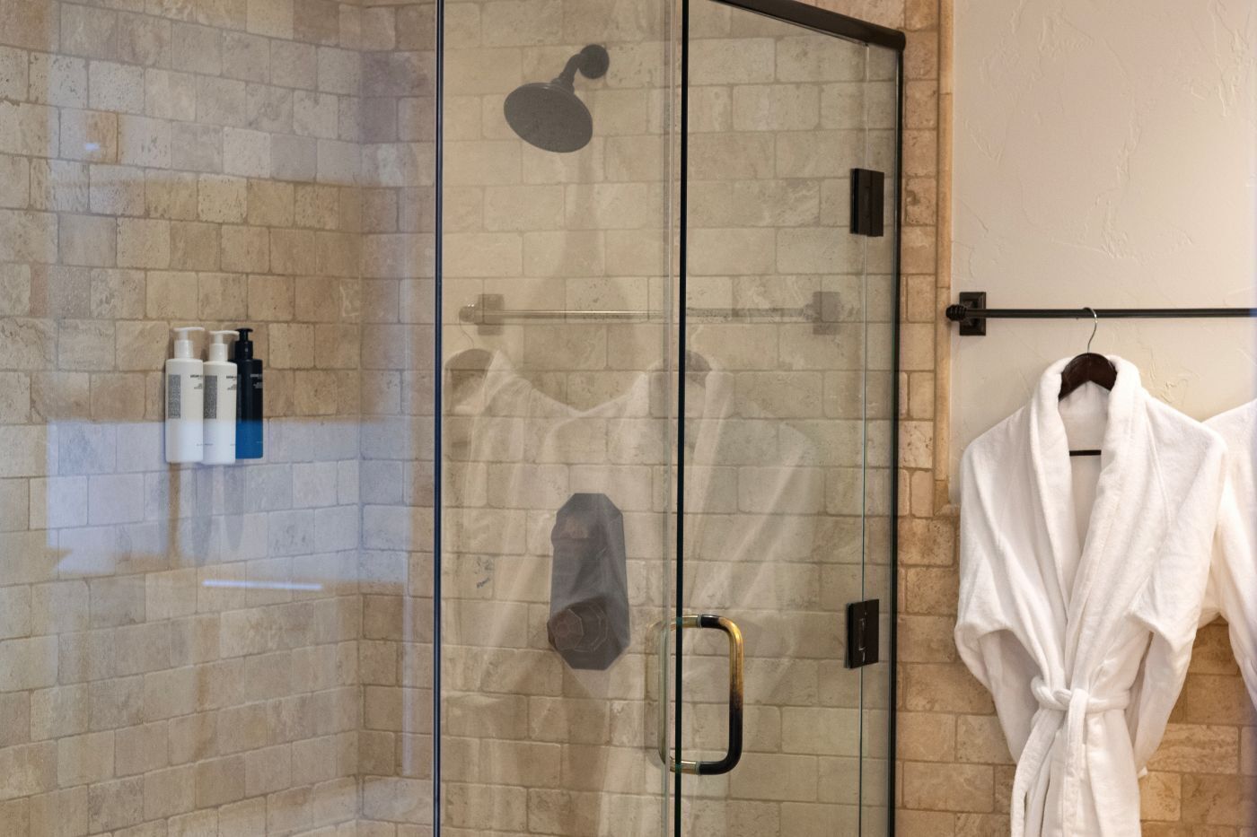 Une douche fermée par des parois en verre, dotée d’un pommeau de douche à main, d’un porte-serviettes avec un peignoir blanc et de produits de toilette fixés au mur.