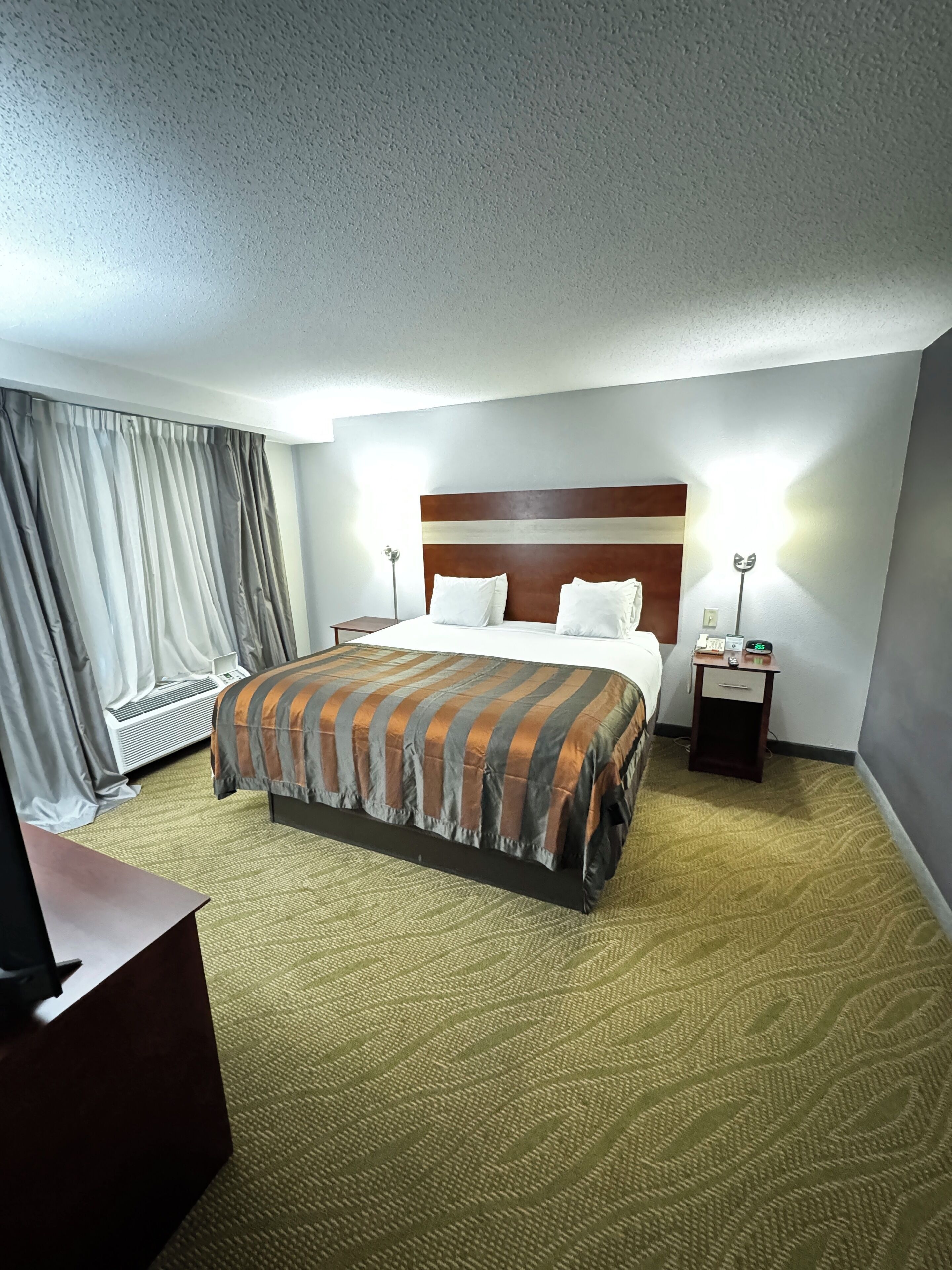 A hotel room with a large bed, a desk, a nightstand, a lamp, and a window with curtains.