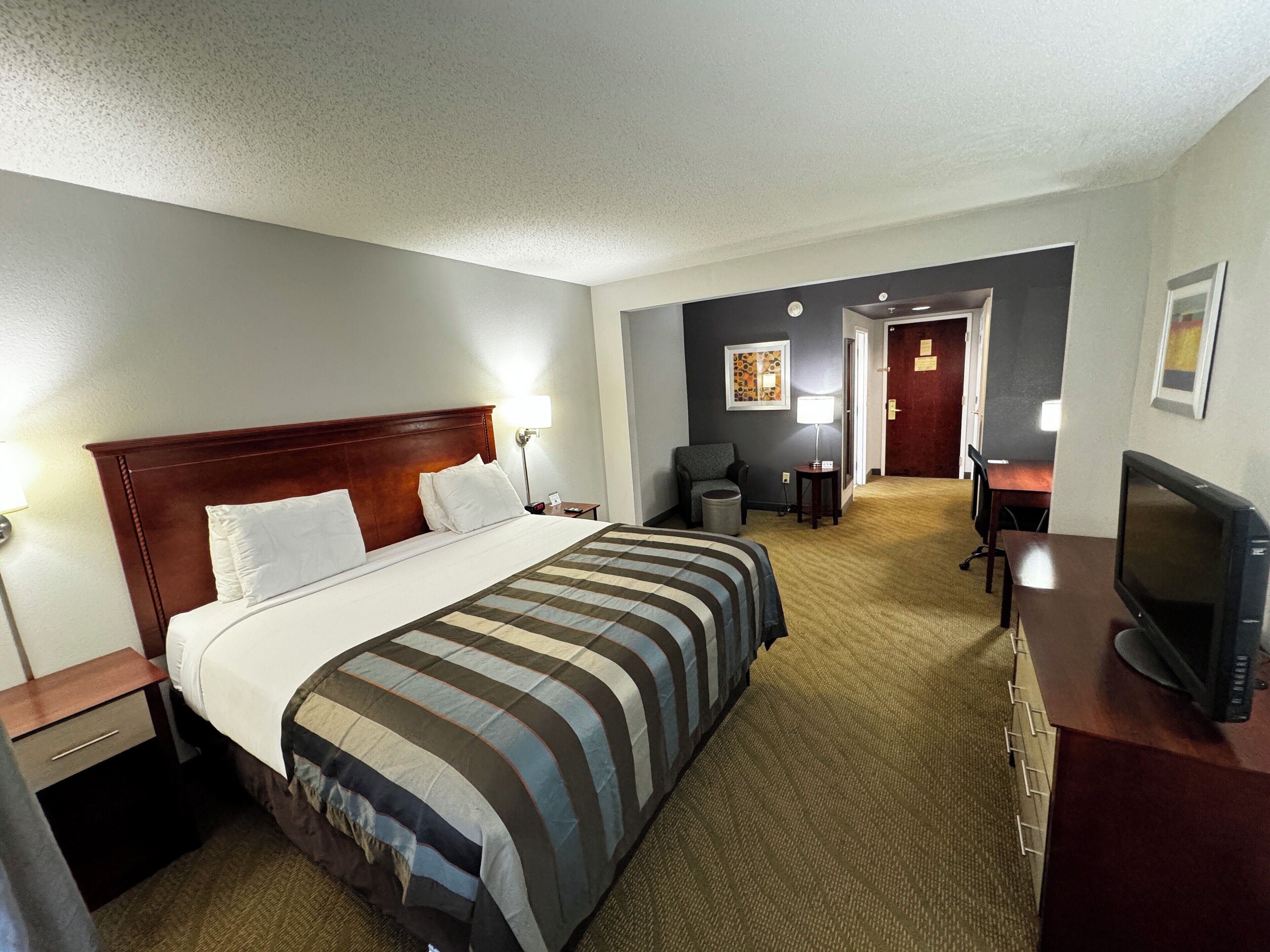 A hotel room with a large bed, a desk with a television, a chair, and a small table with a lamp.
