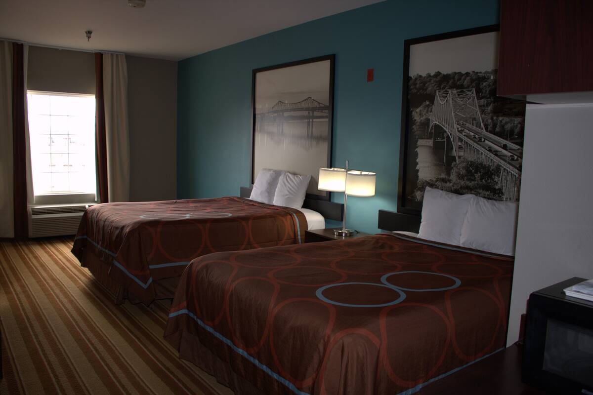 A hotel room with two beds, a large window with curtains, a wall-mounted TV, and a framed picture of a bridge.