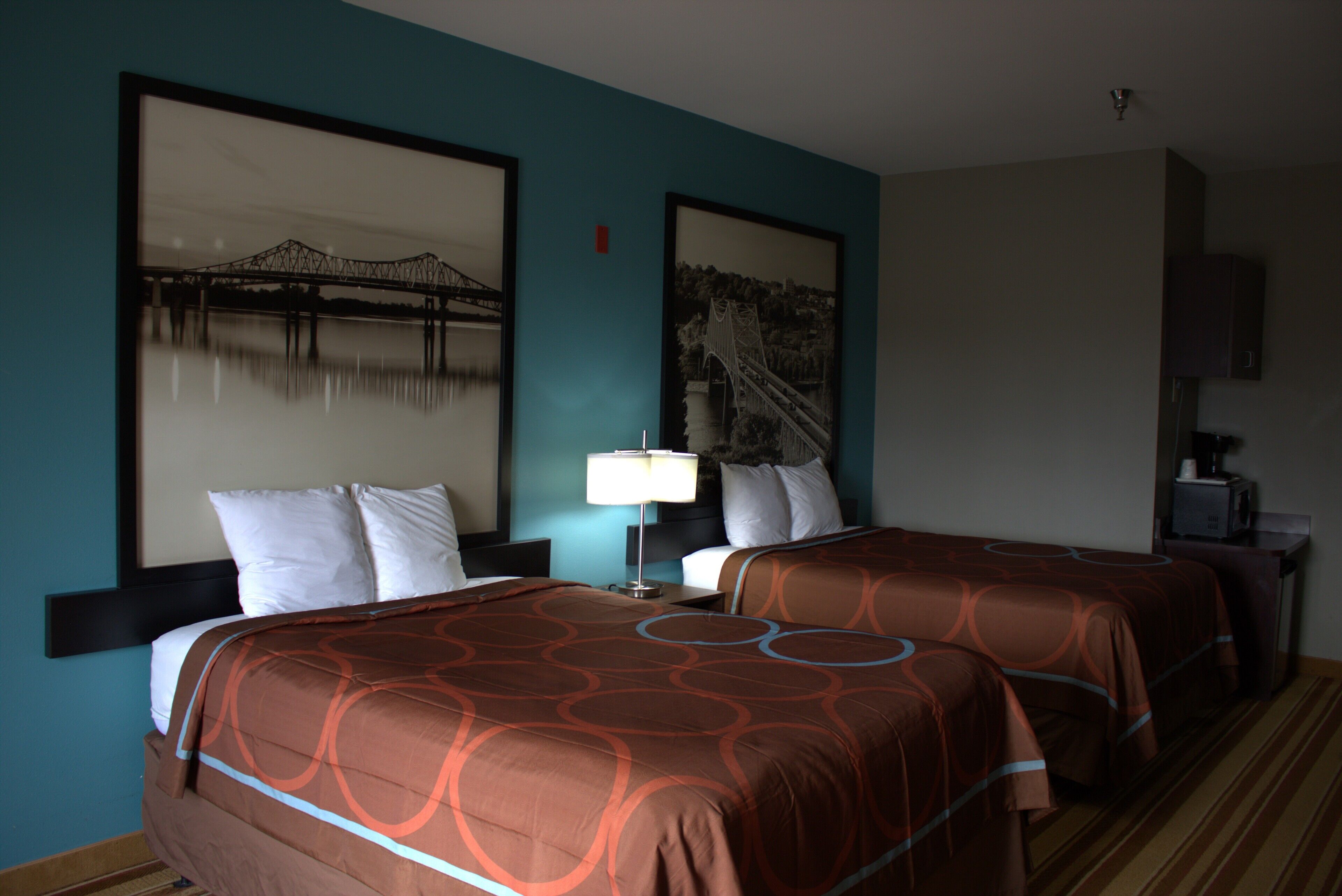 Quarto de hotel com duas camas, uma mesinha de cabeceira com abajur e duas fotografias emolduradas na parede.