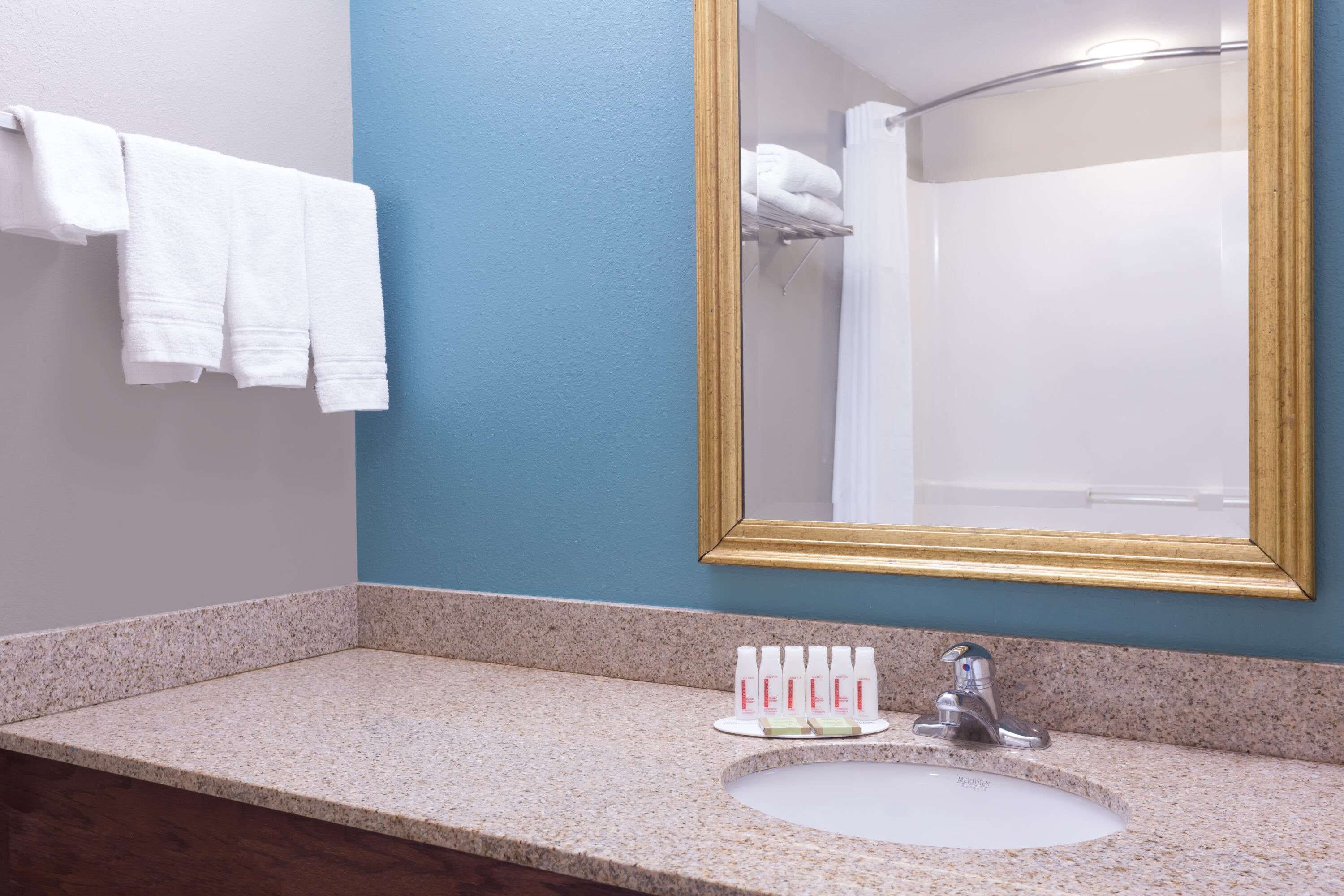 A bathroom with a granite countertop, a white sink, a gold-framed mirror, and a towel rack with white towels.