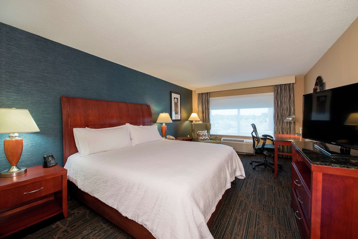 A hotel room with a large bed, two bedside tables with lamps, a desk with a chair, a flat-screen TV, and a window with curtains.