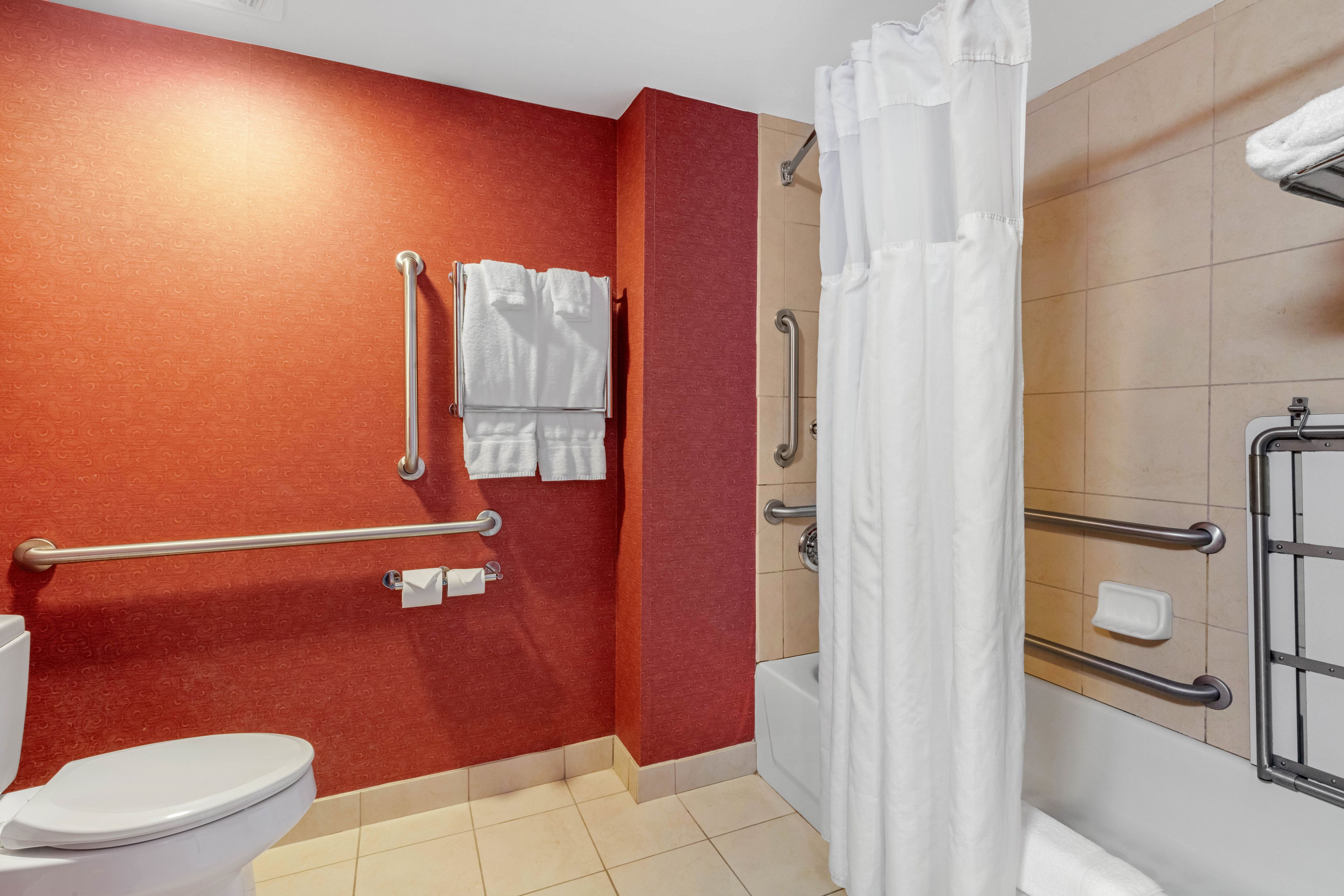 A bathroom with a red wall, a white toilet, a shower with a white curtain, and handrails.
