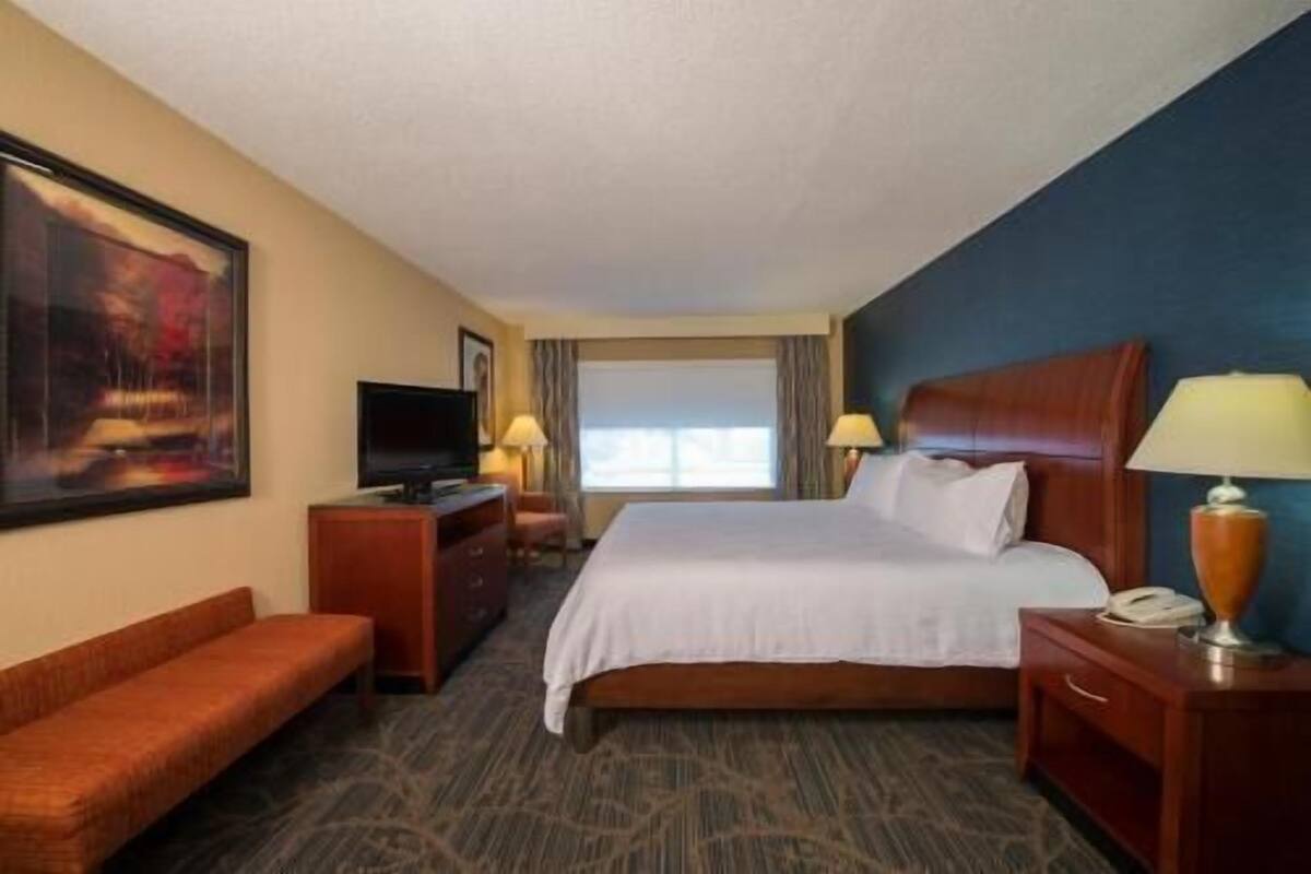 A hotel room with a bed, a desk with a TV, a chair, a bedside table with a lamp, and a painting on the wall.
