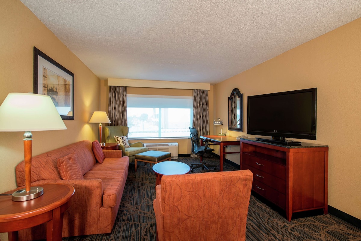 A hotel room with a sofa, armchair, small round table, desk with a TV, and a dresser.