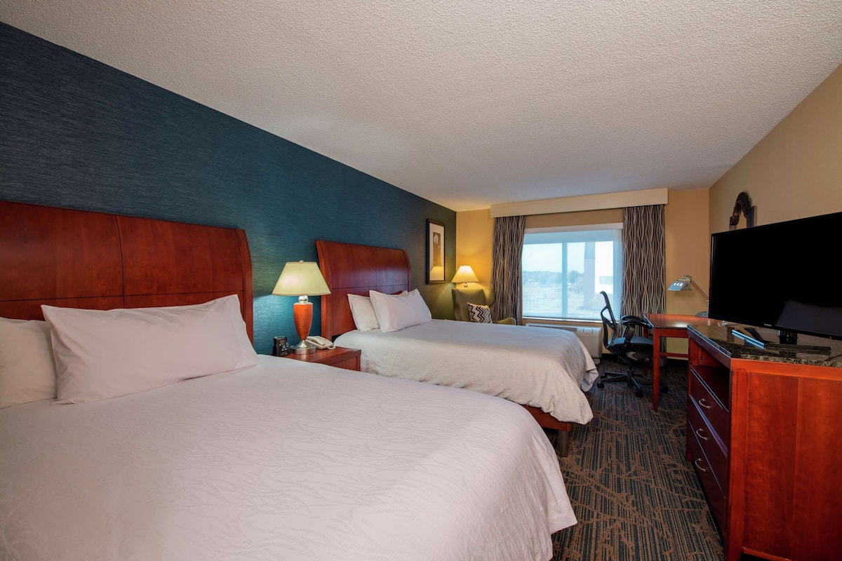 A hotel room with two beds, a desk, a TV, and a window with curtains.