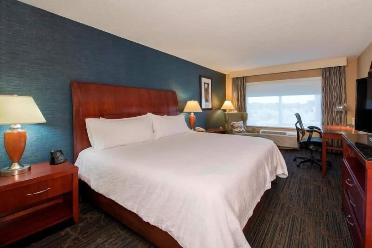 A hotel room with a bed, bedside tables, lamps, a desk with a chair, a television, and a window with curtains.