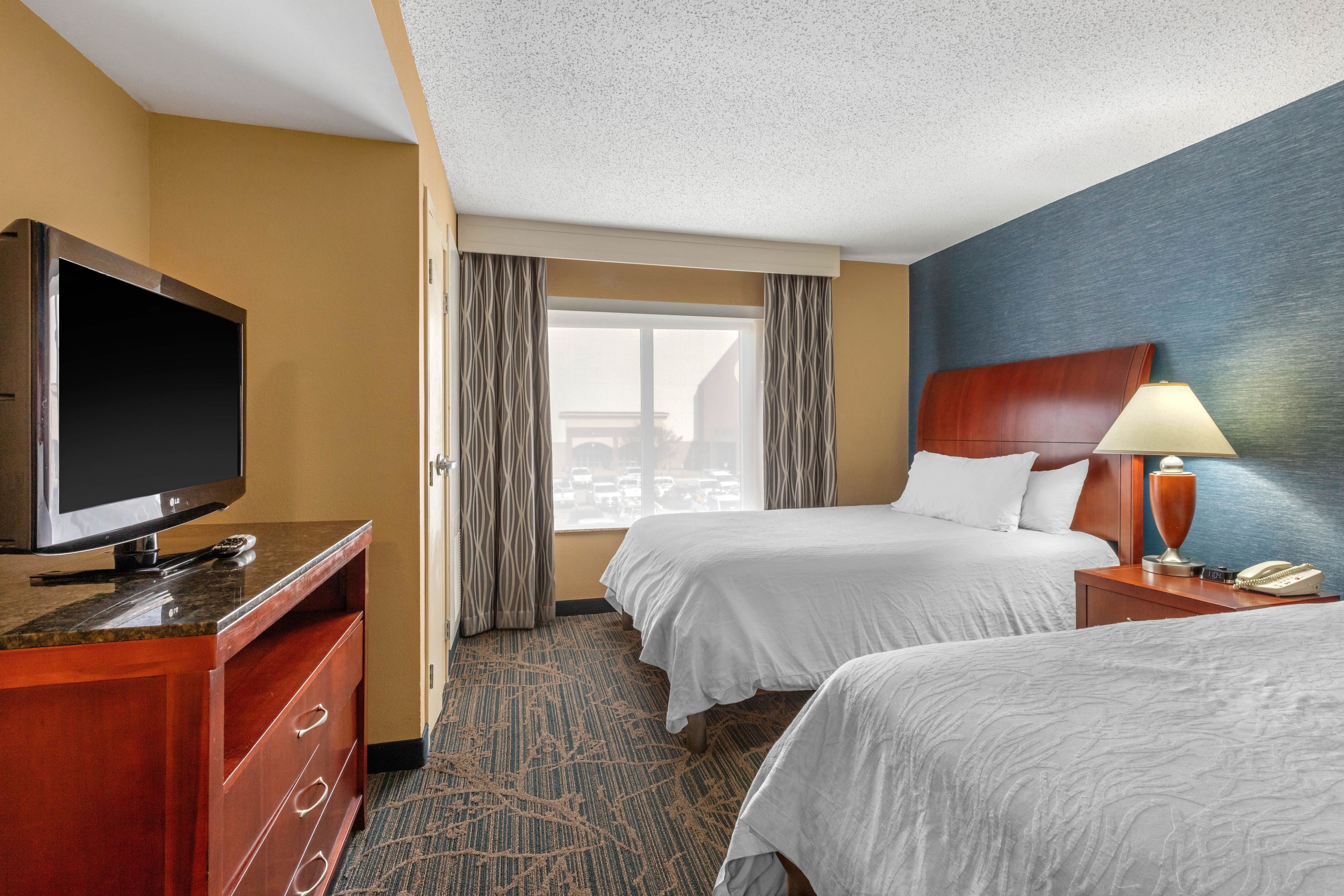 A hotel room with two beds, a TV, a desk, and a window with curtains.