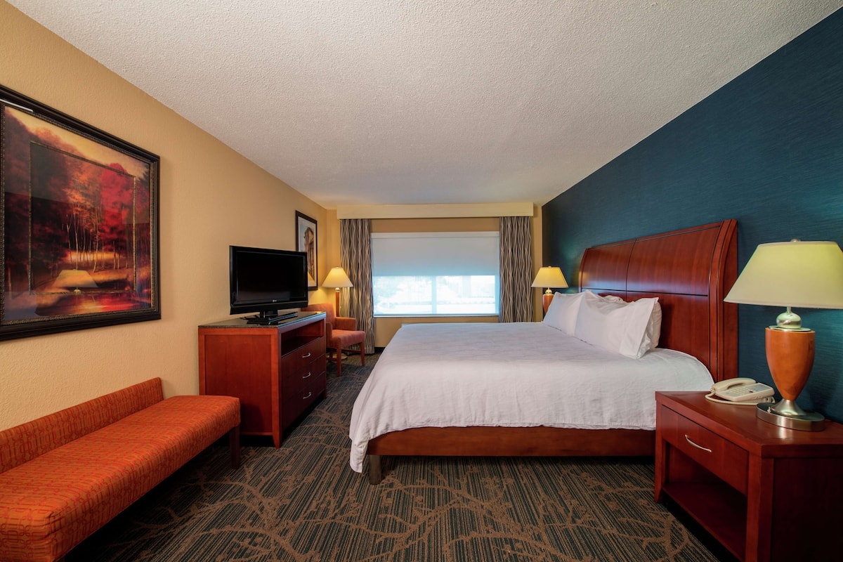 A hotel room with a large bed, a TV, a desk with a lamp, a bench, and a painting on the wall.