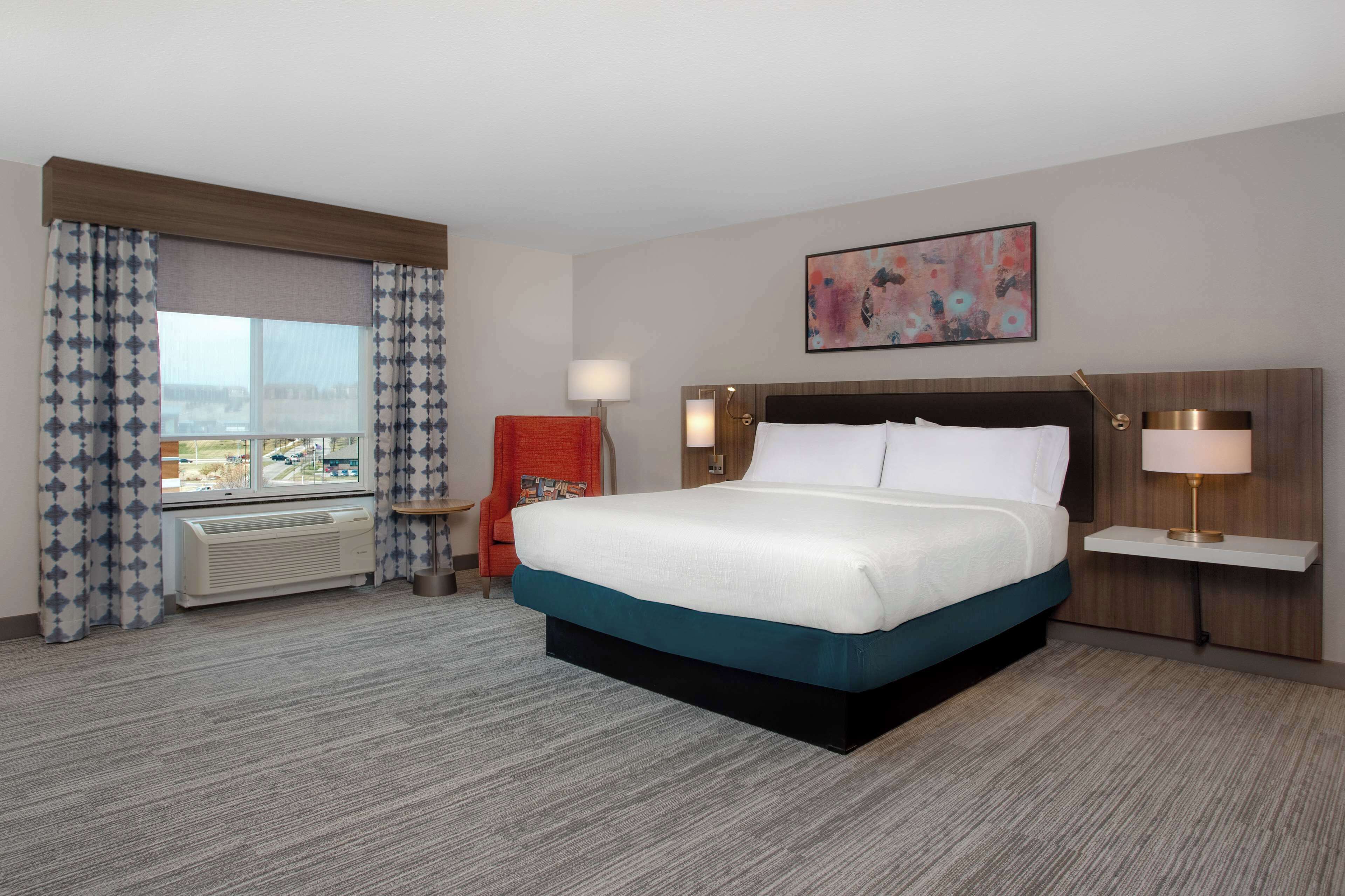 Habitación de hotel con una cama grande, una silla, una mesita de noche, una lámpara y una ventana con cortinas.