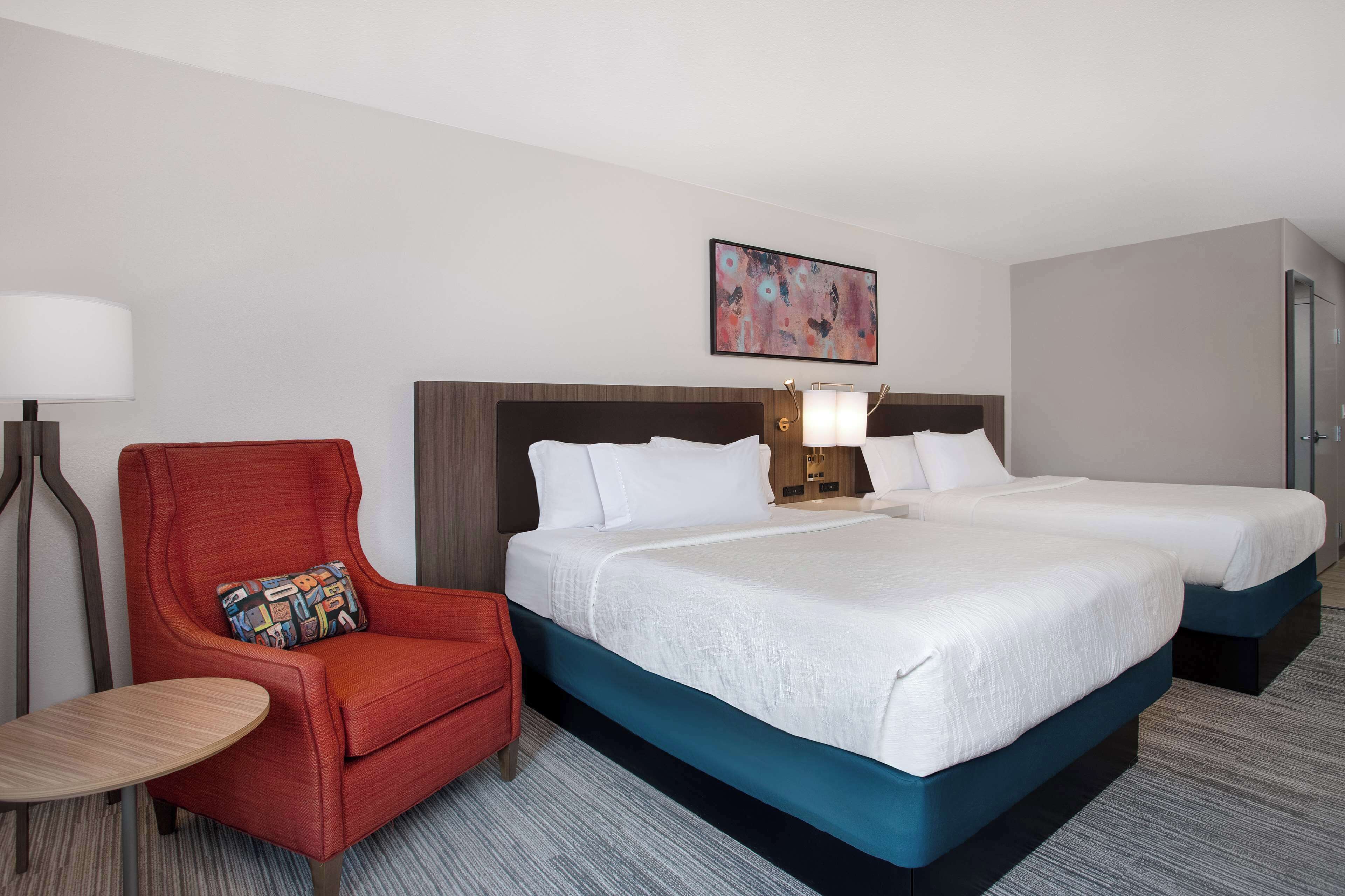A hotel room with two beds, a red armchair, a small table, and a painting on the wall.