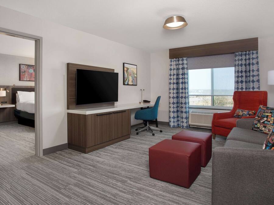 Une chambre d’hôtel équipée d’une télévision à écran plat, d’un bureau avec une lampe, d’un fauteuil de bureau bleu, d’un fauteuil rouge et d’un canapé gris agrémenté de coussins à motifs.