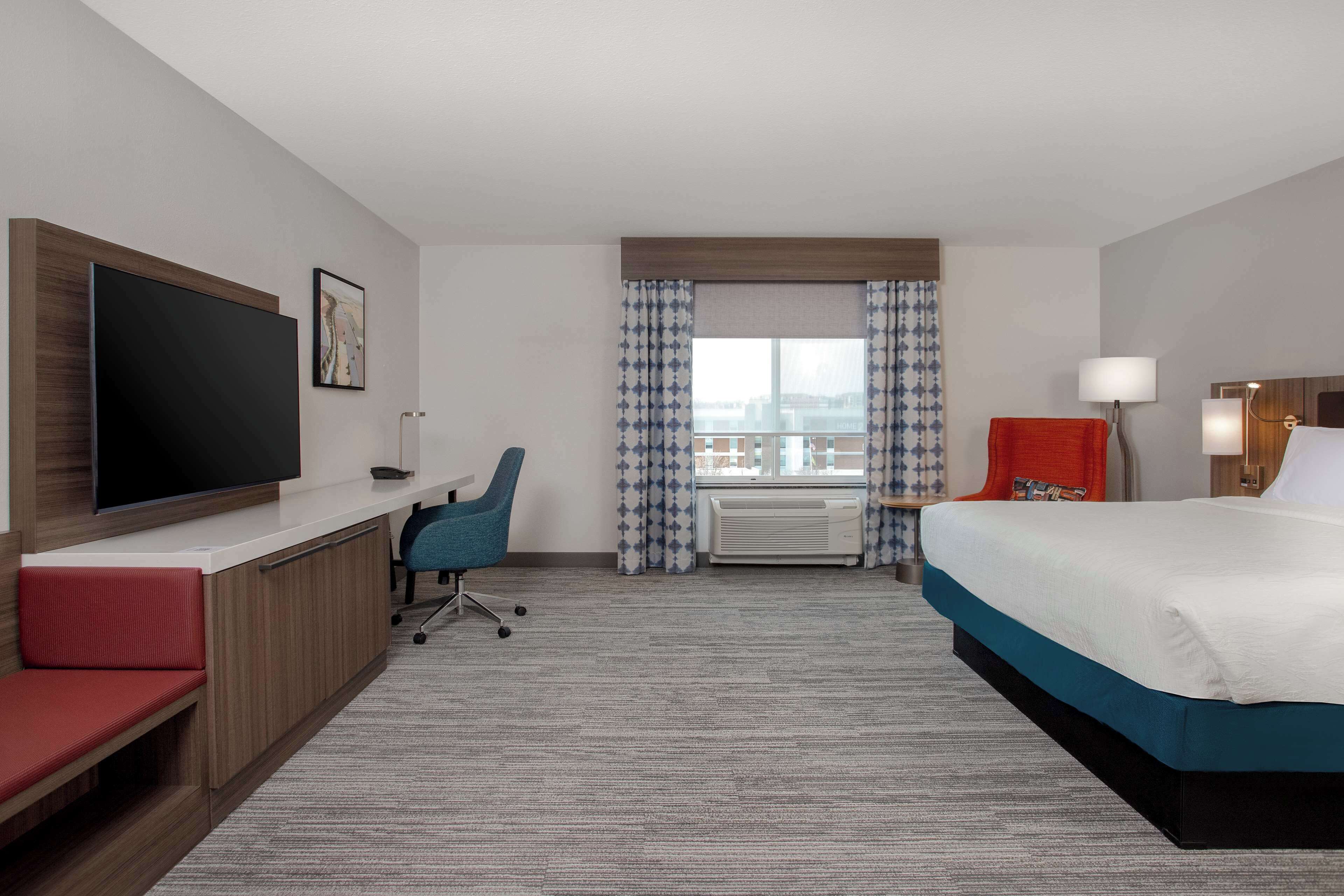 Habitación de hotel con cama, escritorio, televisor, silla y ventana con cortinas.