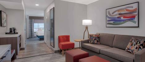 Une chambre d’hôtel moderne avec un canapé gris, un pouf rouge et une petite table avec une lampe. Un tableau abstrait aux couleurs vives est accroché au mur, et on aperçoit une salle de bain au fond.