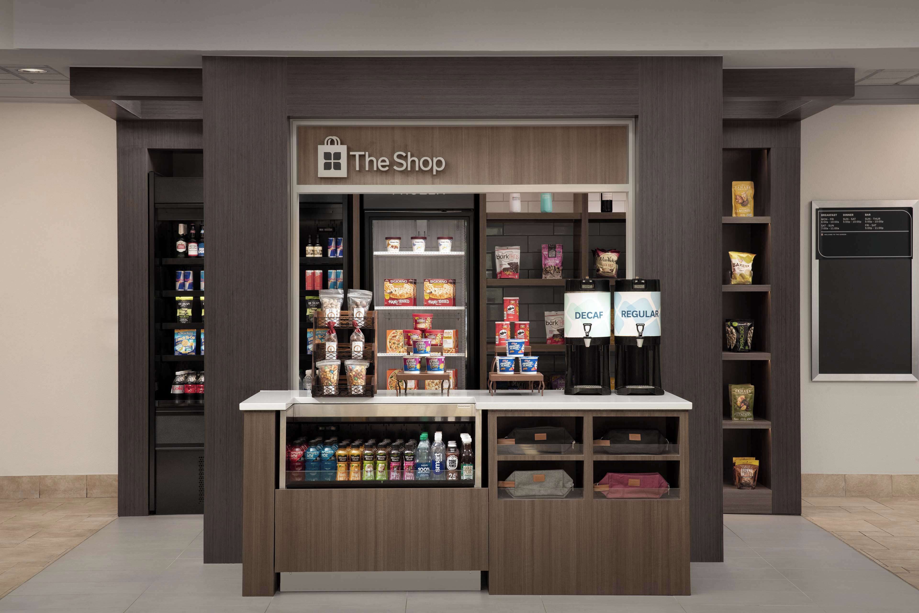 Un moderno quiosco comercial con un letrero que dice "The Shop", que presenta una máquina de café, estantes con aperitivos y una variedad de productos envasados.