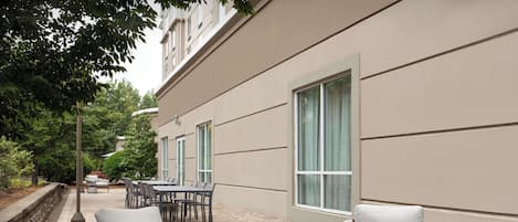 An outdoor patio with wicker chairs and a round table, adjacent to a building with large windows and a stone wall.
