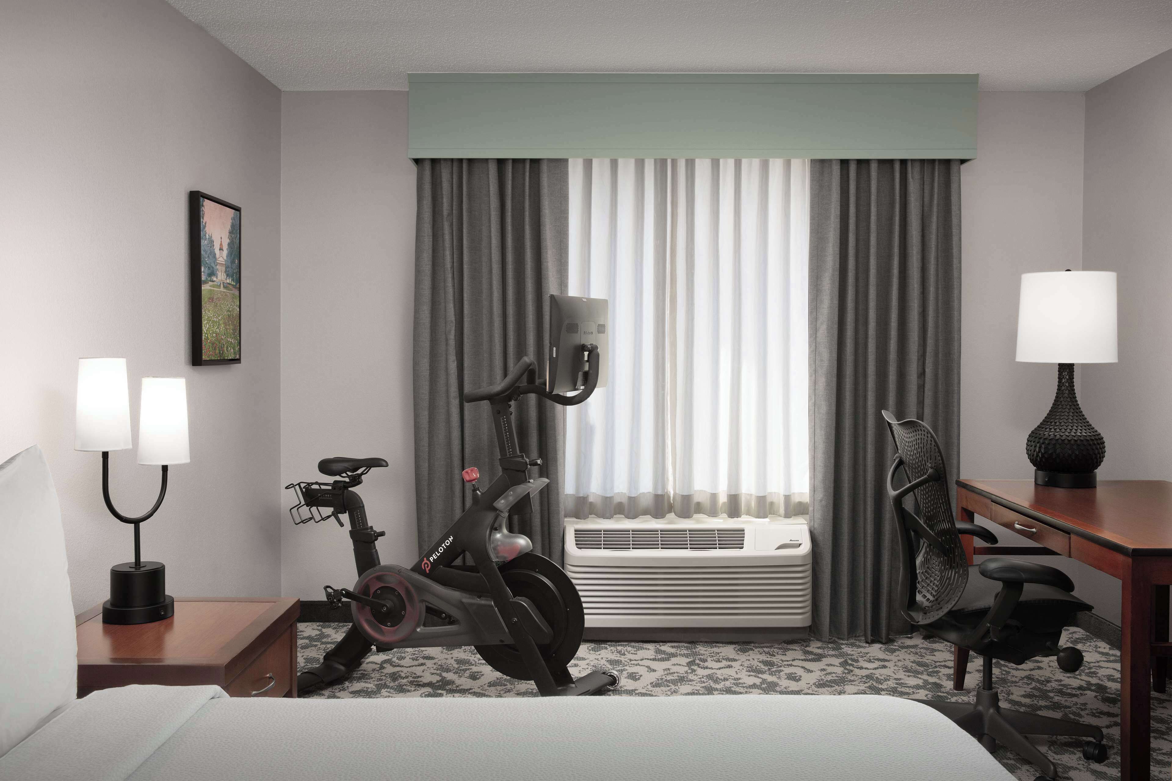 A hotel room with a bed, a desk with a lamp, an exercise bike, a chair, and a window with curtains.