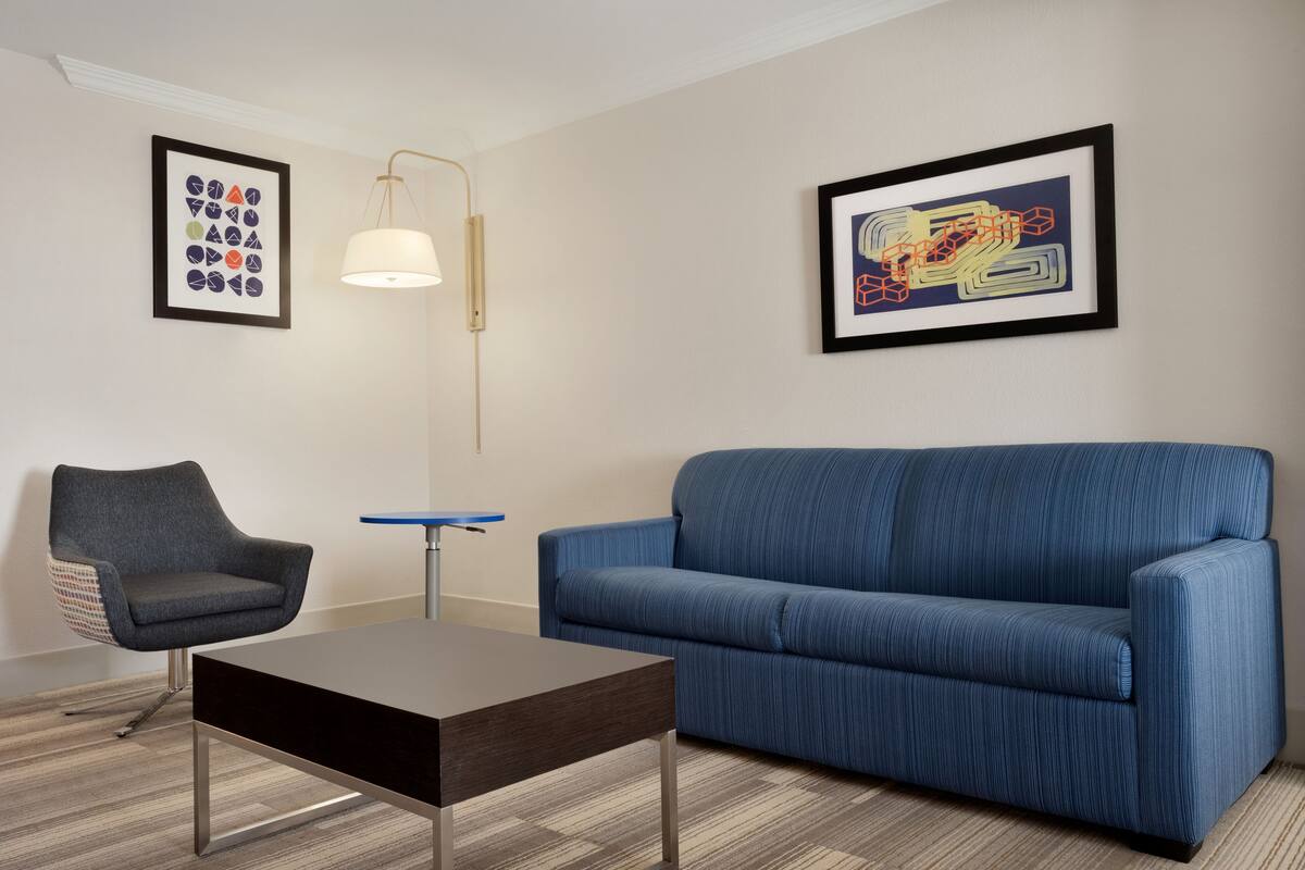A modern living room with a blue sofa, a dark wooden coffee table, a grey armchair, and a floor lamp. Two framed artworks are hung on the wall.